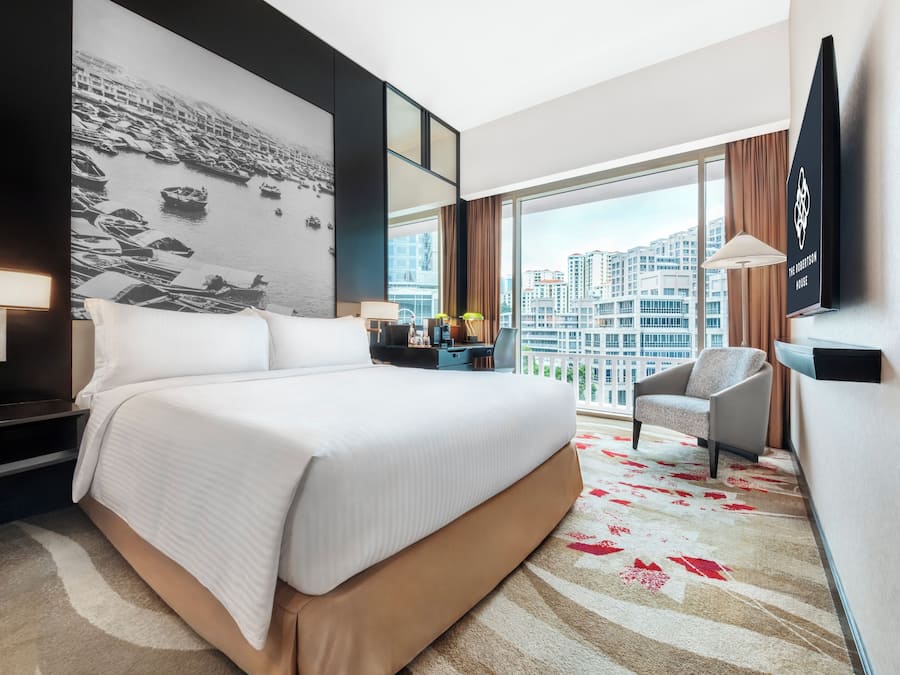 Habitación de hotel con una cama grande, un escritorio, una silla, un televisor y vistas de la ciudad a través de amplios ventanales.