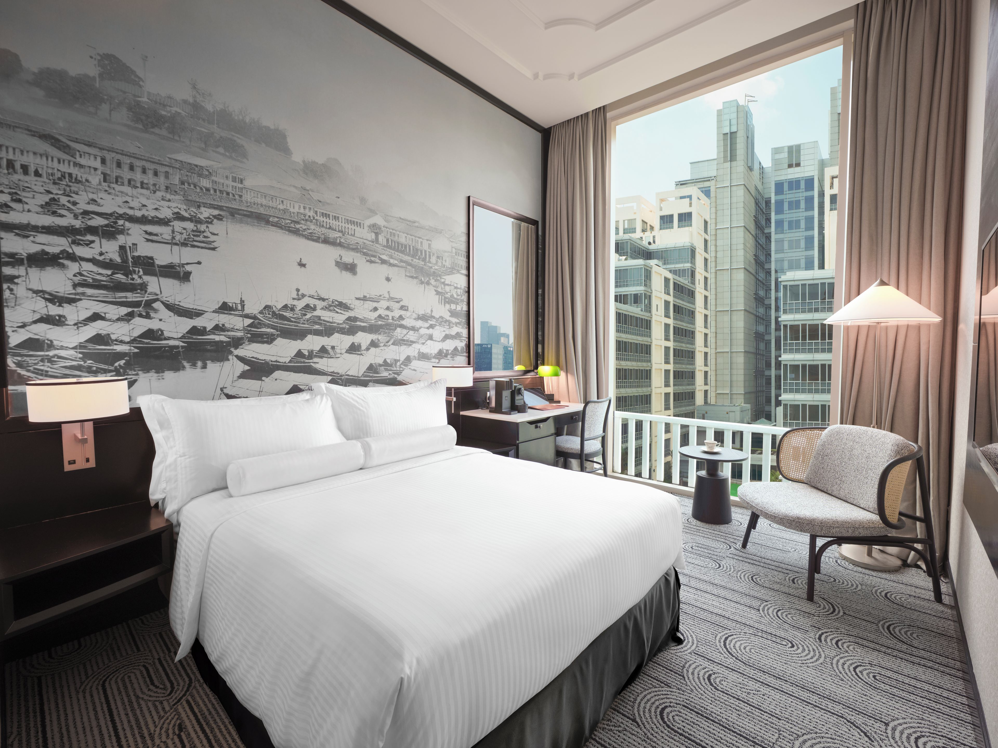 A hotel room with a large bed, a desk, a chair, a view of tall buildings, and a wall with a black and white photo.