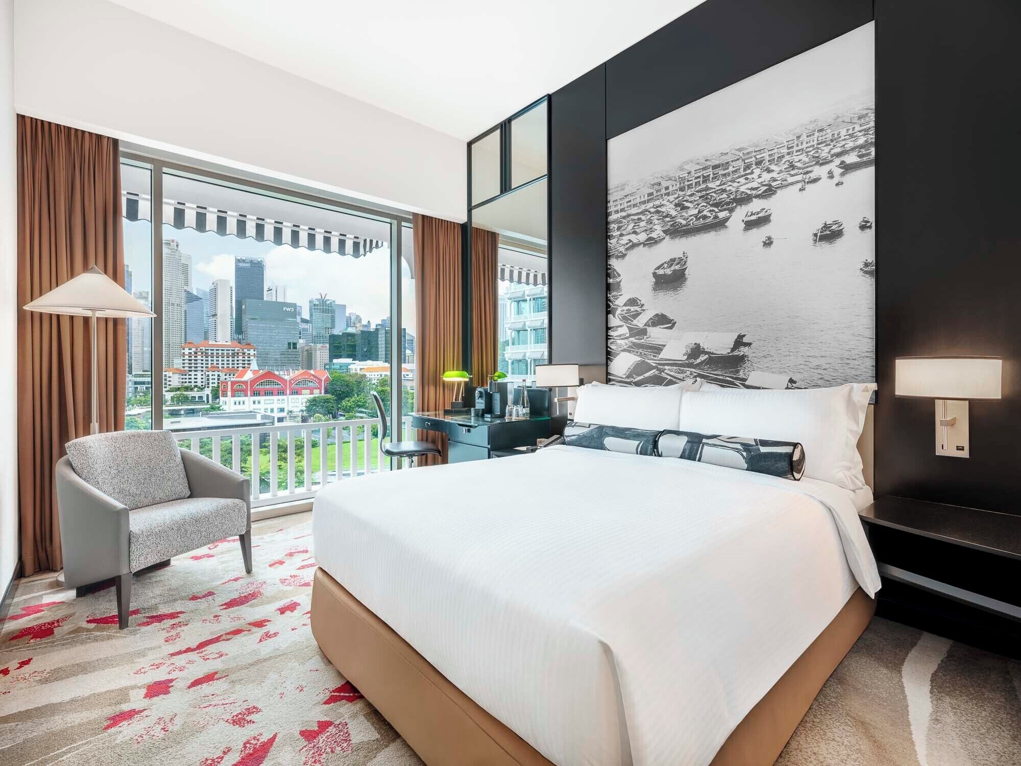 Habitación de hotel con una cama grande, una silla, un escritorio, vista a la ciudad y una pared con una fotografía blanco y negro.