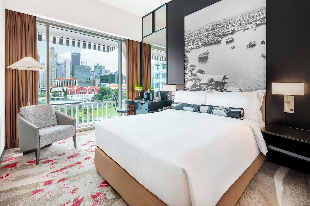 Habitación de hotel con una cama grande, una silla, un escritorio, vista a la ciudad y una pared con una fotografía blanco y negro.