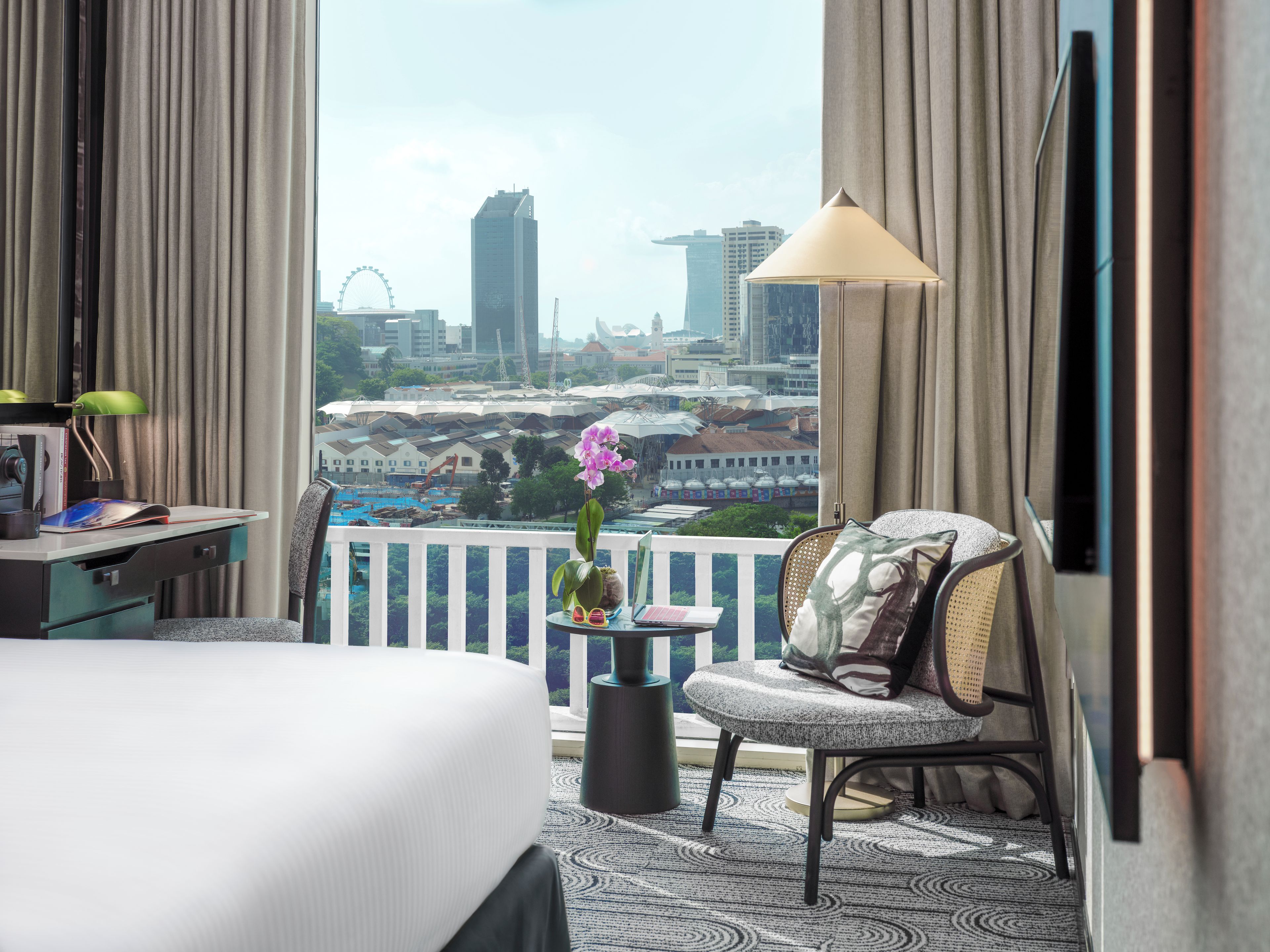 Habitación de hotel con cama, escritorio, silla, una pequeña mesa con un jarrón de flores y vistas a un paisaje urbano con edificios y una noria.