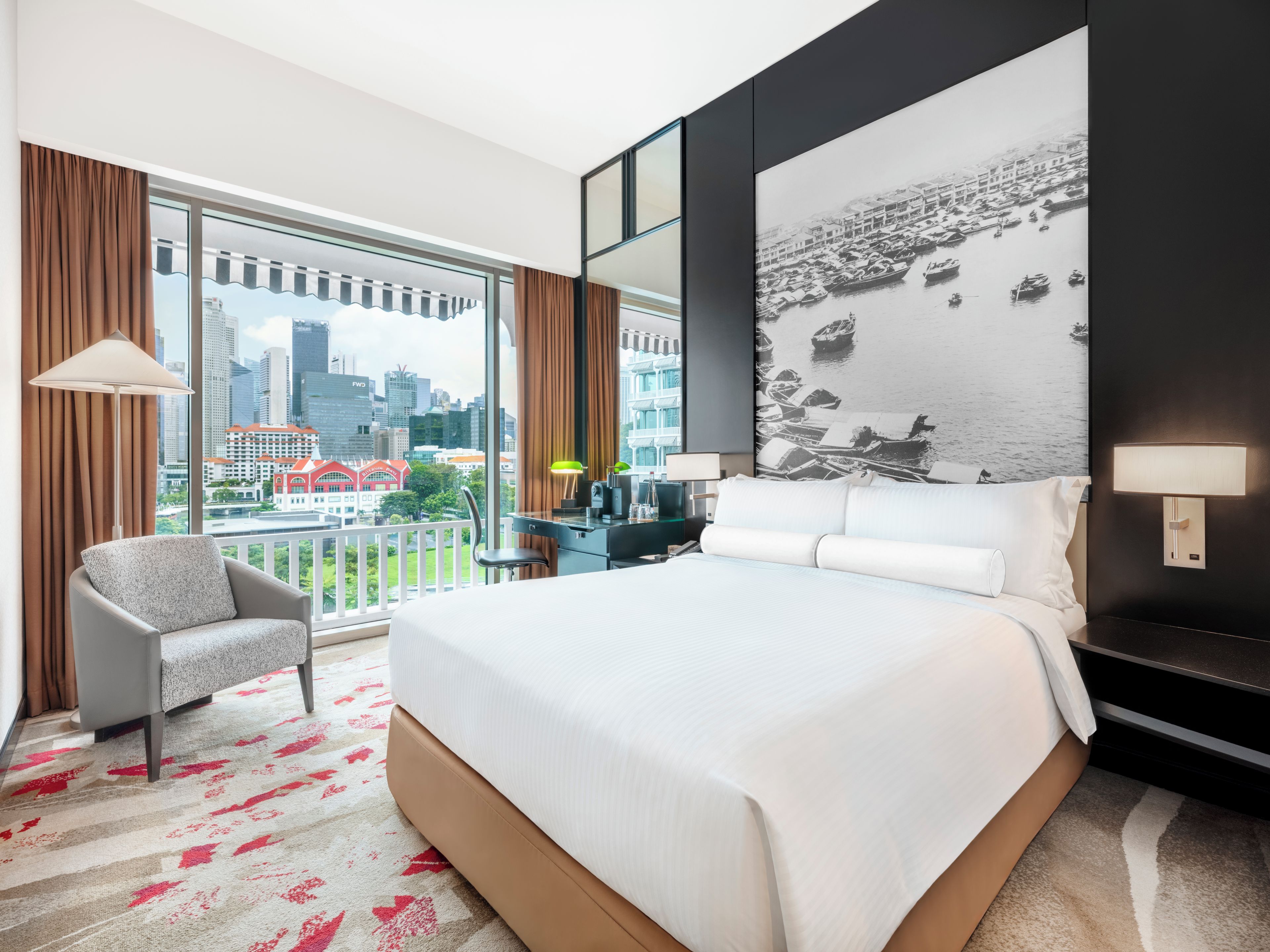 A hotel room with a large bed, a chair, a desk, a city view, and a wall-mounted lamp.