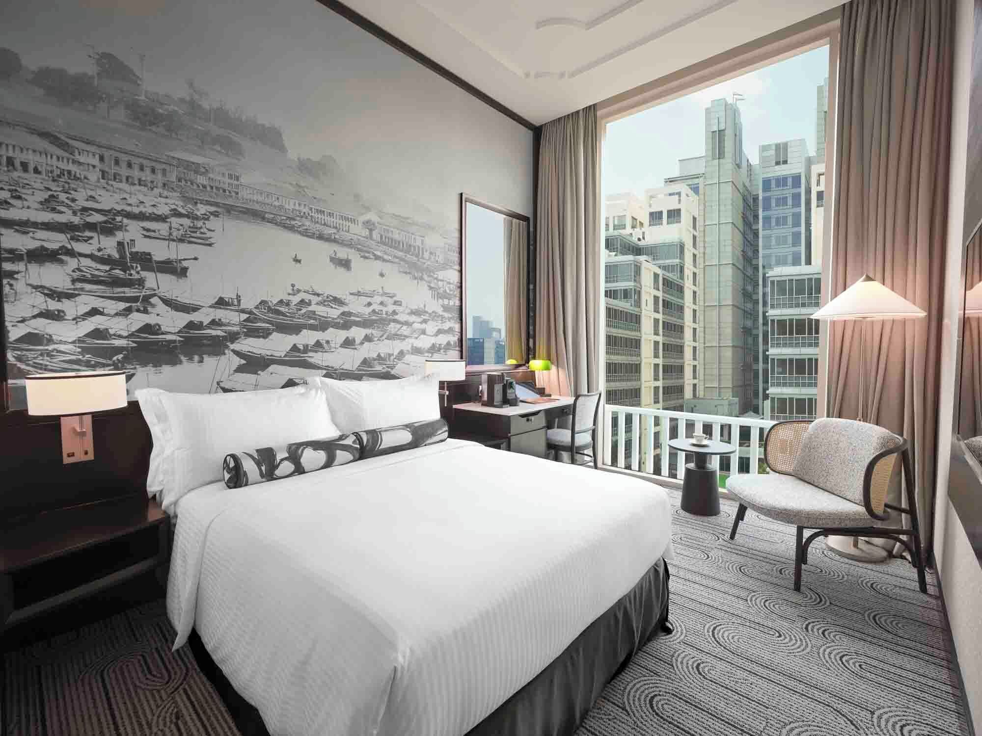 Habitación de hotel con una cama grande, un escritorio, una silla, un balcón con vistas a edificios y un mural en la pared.