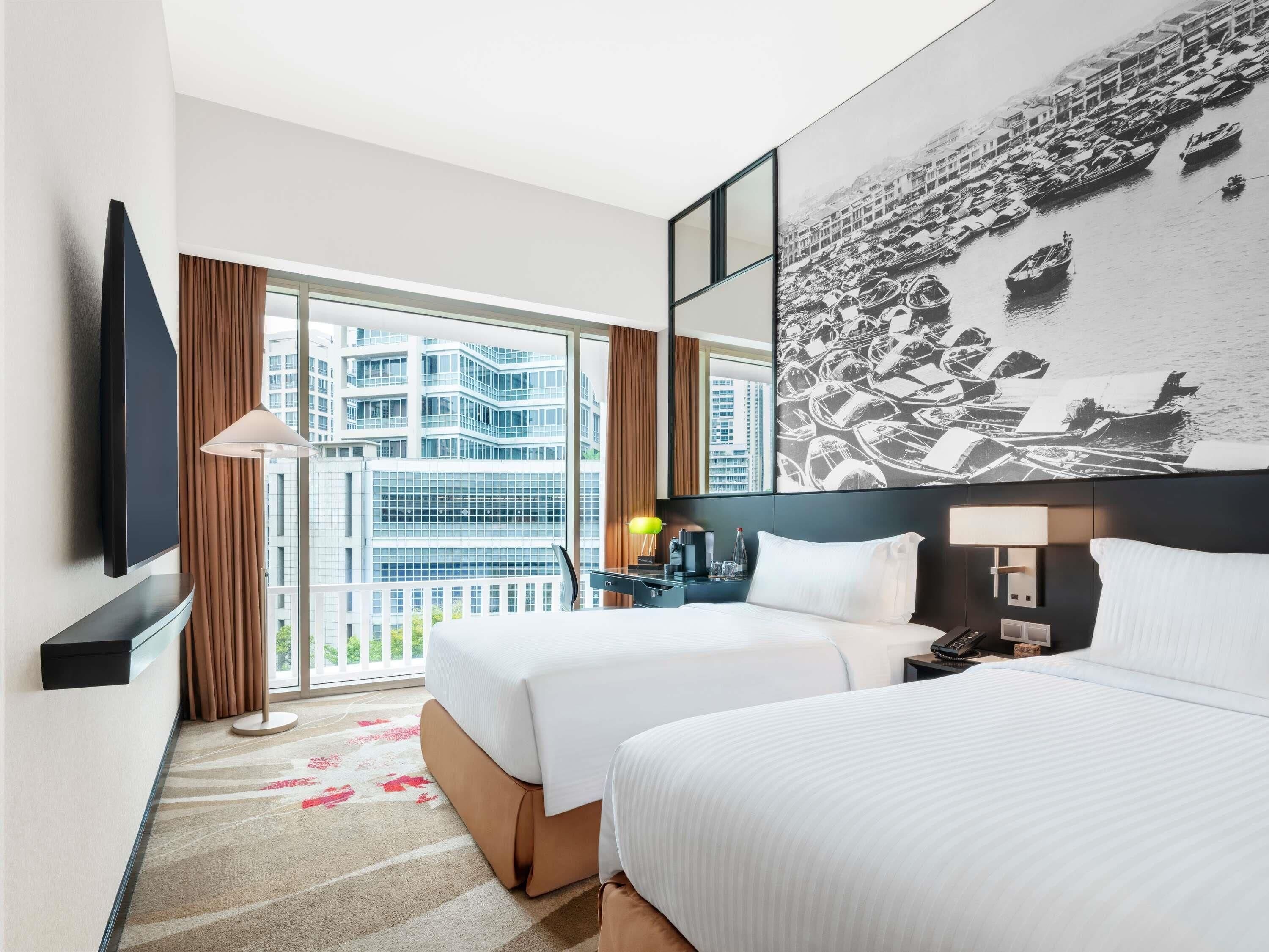 Ein Hotelzimmer mit zwei Betten, einem großen Fenster mit Blick auf Gebäude, einem Flachbildfernseher und einem an der Wand befestigten Kunstwerk.