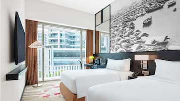 Habitación de hotel con dos camas, un ventanal con vistas a edificios, un televisor de pantalla plana y una obra de arte colgada en la pared.