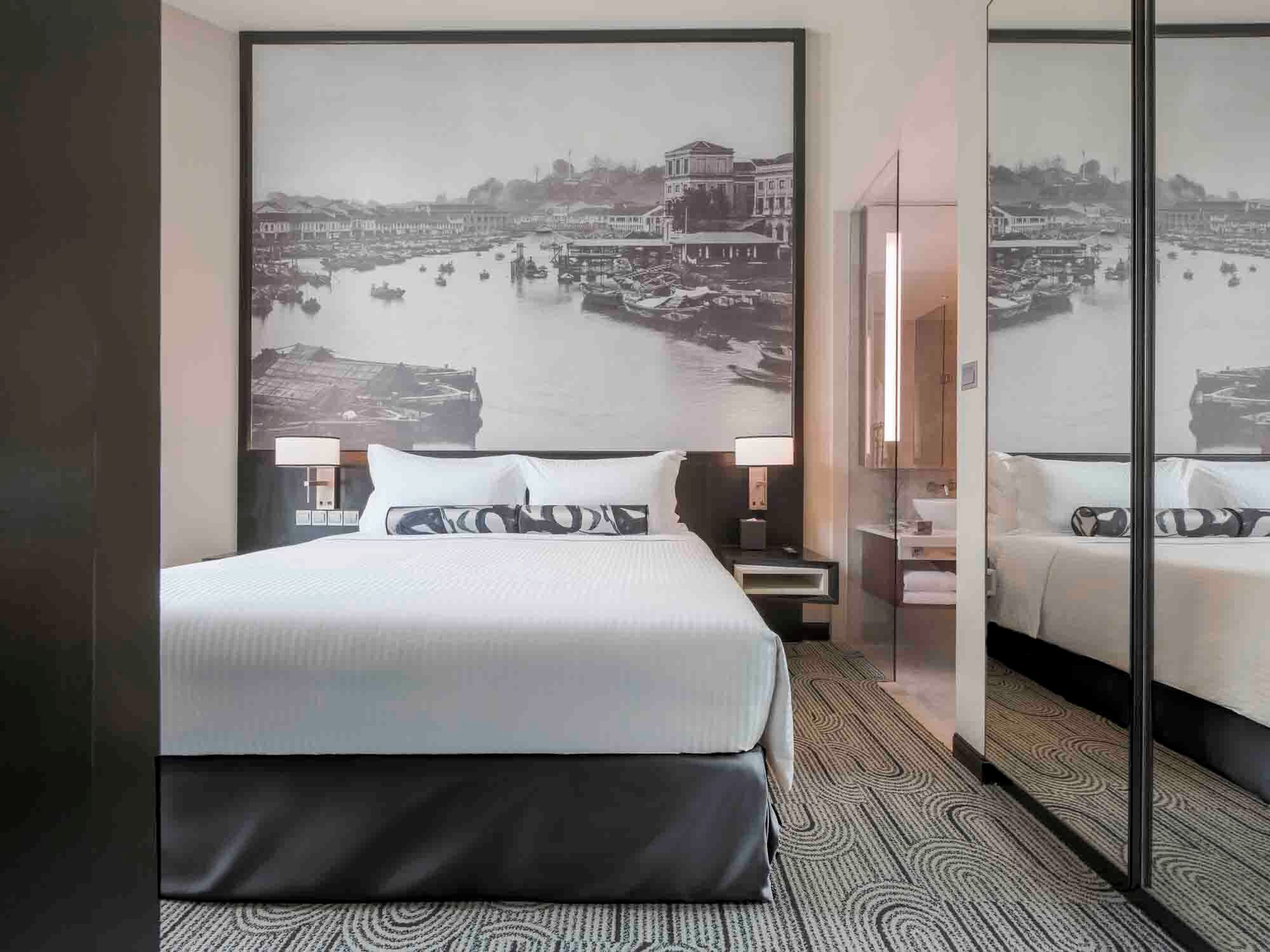 Habitación de hotel con una cama grande, una mesita de noche, un espejo y un cuadro colgado en la pared que muestra una vista acuática.