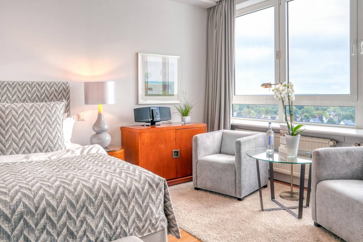 A modern hotel room with a large bed, a sofa, a glass coffee table, a wooden cabinet, and a view of the city through large windows.