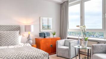 A modern hotel room with a large bed, a sofa, a glass coffee table, a wooden cabinet, and a view of the city through large windows.