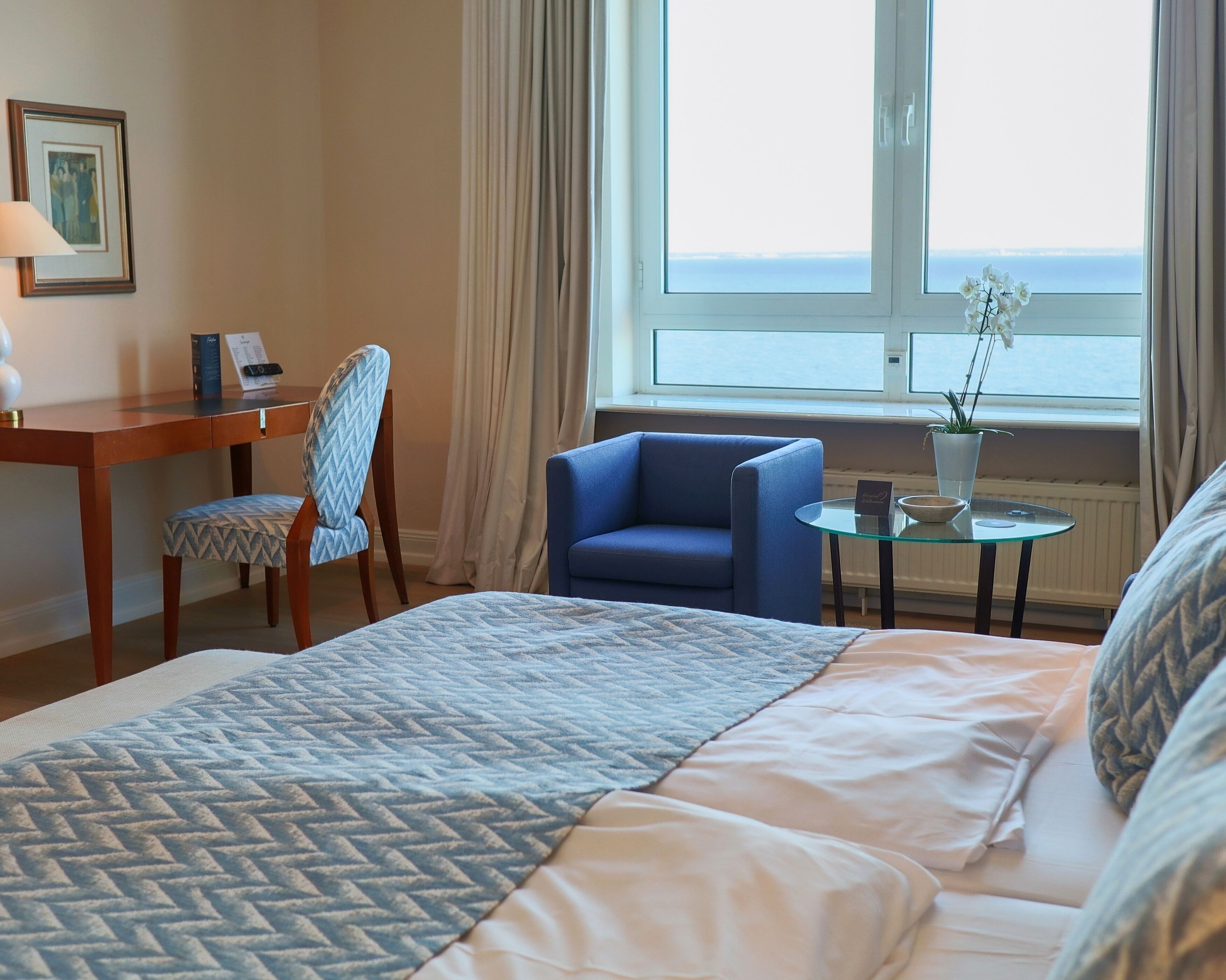 Una habitación de hotel con cama, escritorio, silla, una pequeña mesa con una planta y vista al mar por la ventana.