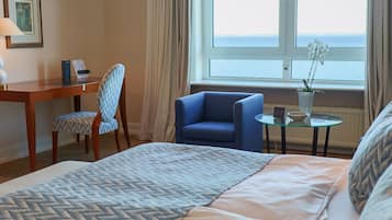 Ein Hotelzimmer mit einem Bett, einem Schreibtisch, einem Stuhl, einem kleinen Tisch mit einer Pflanze und Meerblick durch das Fenster.