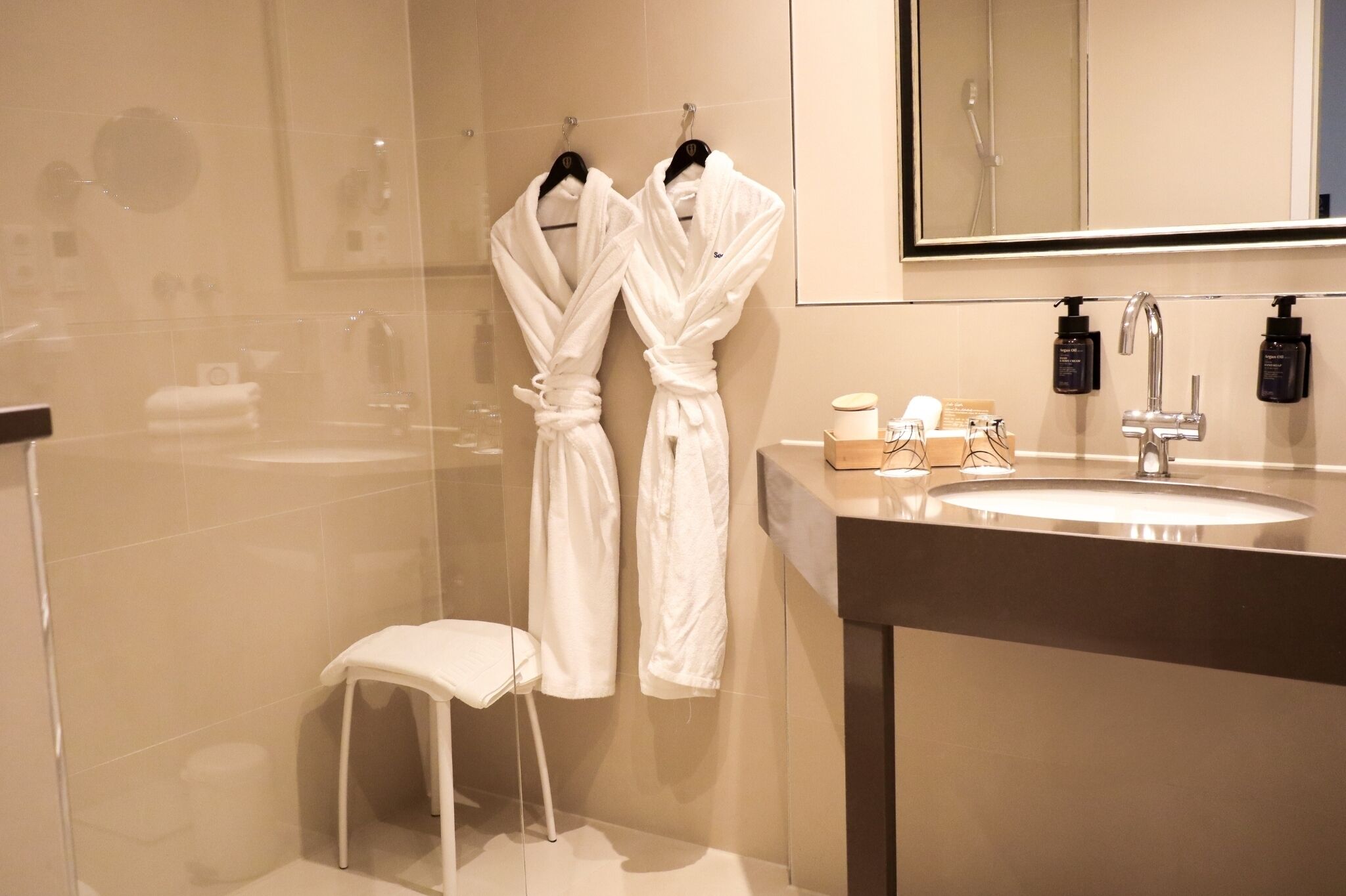 A modern bathroom with a glass shower enclosure, a white sink with a mirror above it, and two white robes hanging on hooks.