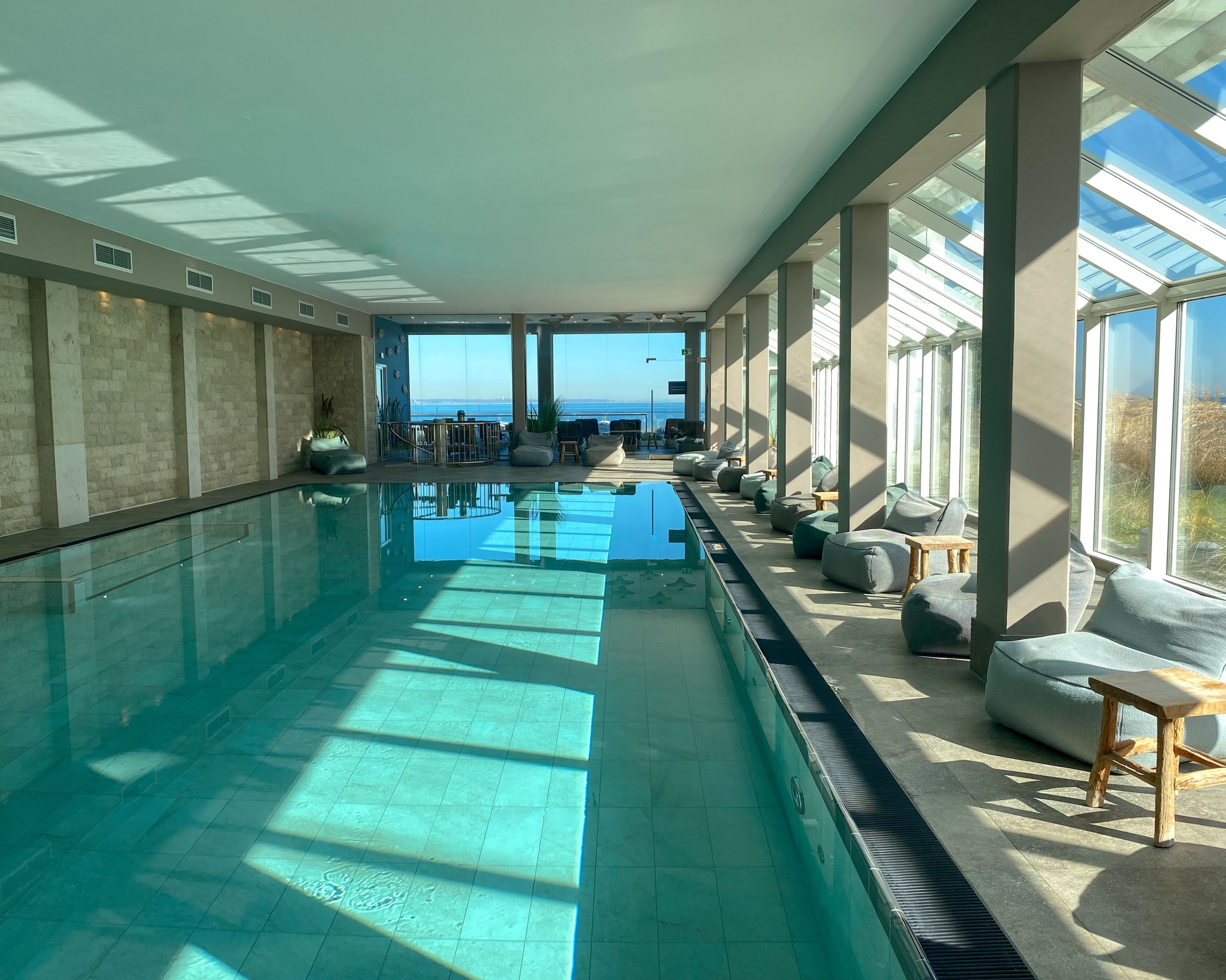 Una piscina interior larga con vista al mar, rodeada de amplios ventanales y zonas de estar.