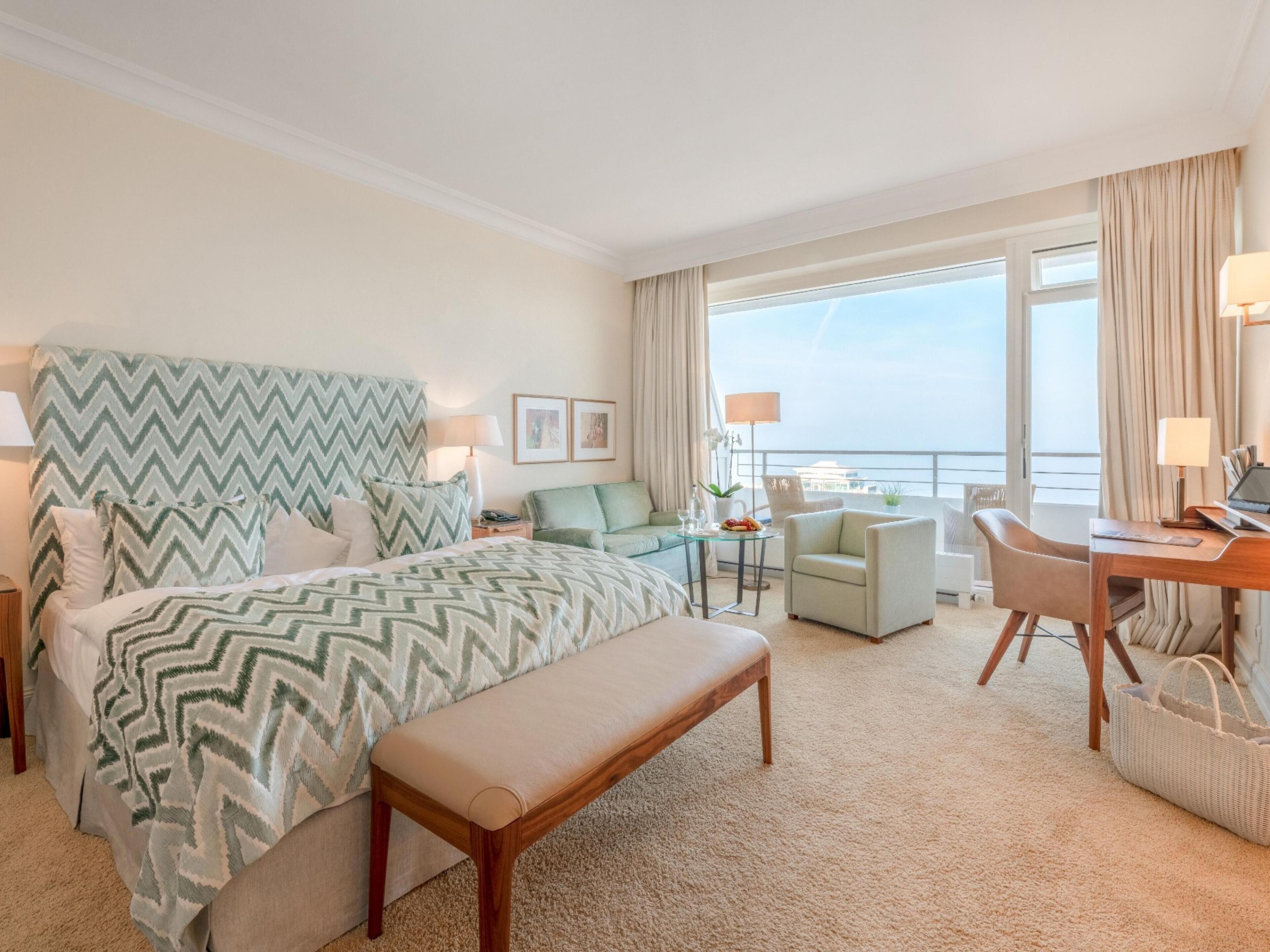 Habitación de hotel con cama, escritorio, silla, banca, mesita y vista al mar.