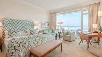 Habitación de hotel con cama, escritorio, silla, banca, mesita y vista al mar.