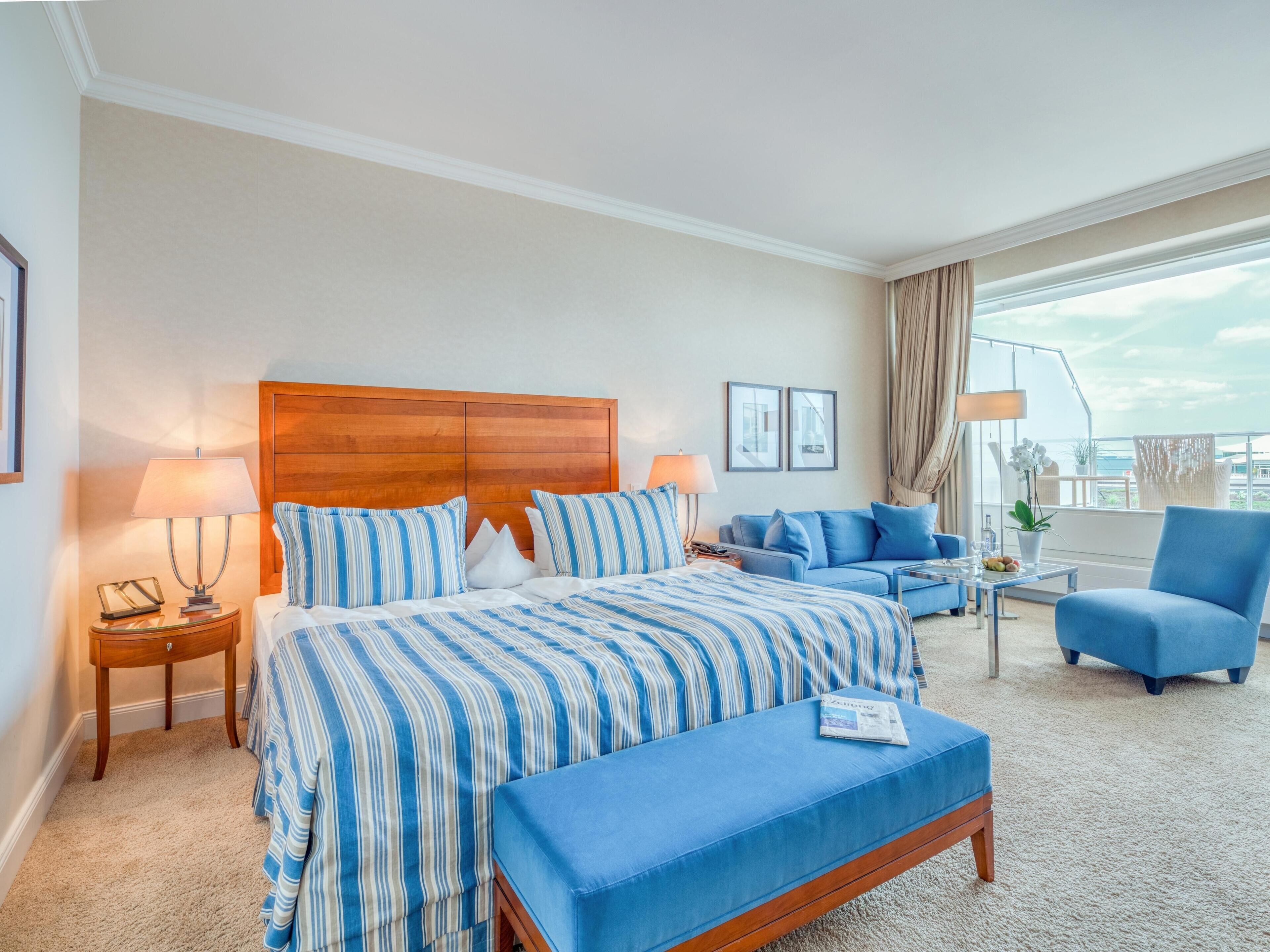 A hotel room with a large bed, a sofa, a chair, and a balcony with a view.