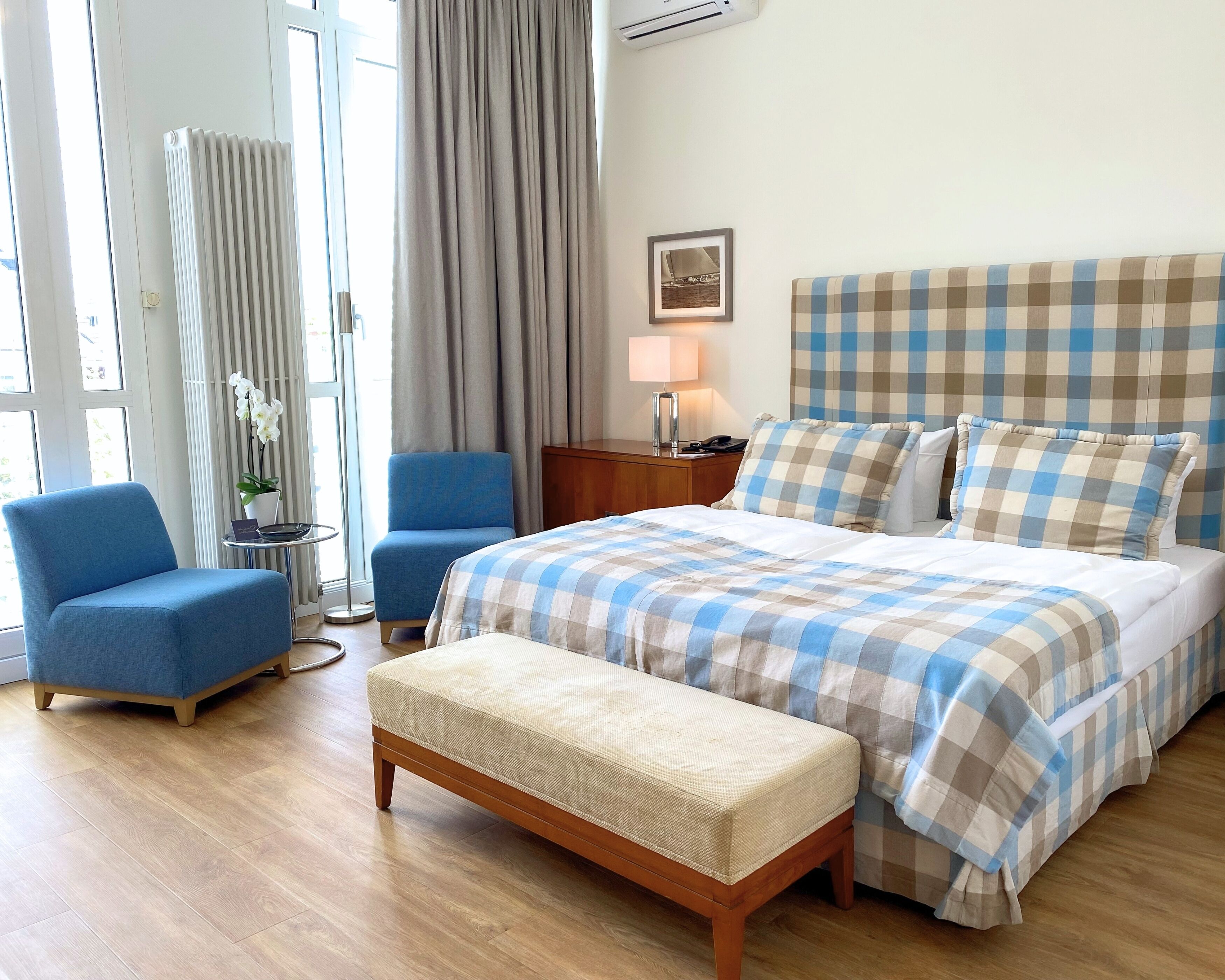 Une chambre d’hôtel avec un lit, un fauteuil bleu, un banc en bois, une petite table avec une lampe et une fenêtre avec des rideaux.