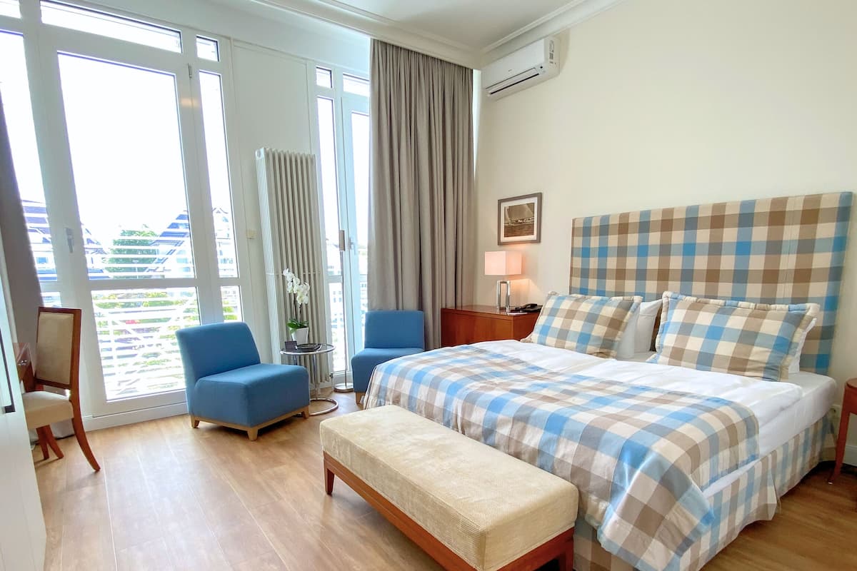 A hotel room with a large bed, a bench, two armchairs, a small table with a lamp, and a view through the glass doors.