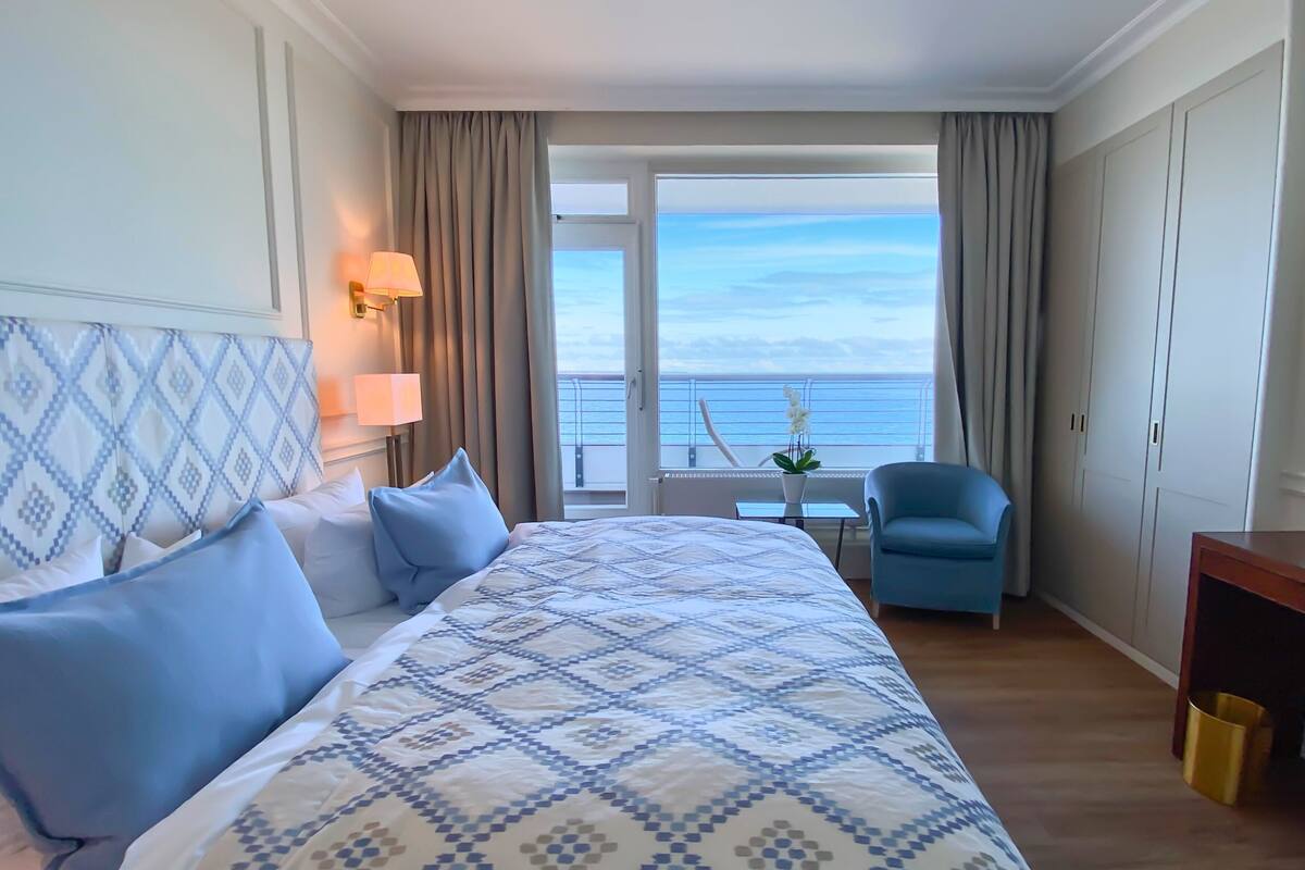 A hotel room with a large bed, a chair, a small table, and a view of the sea.
