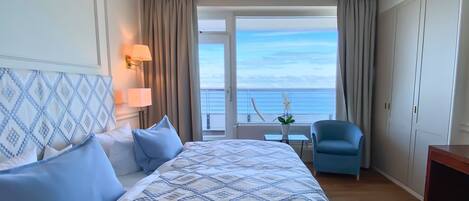 A hotel room with a large bed, a chair, a small table, and a view of the sea.