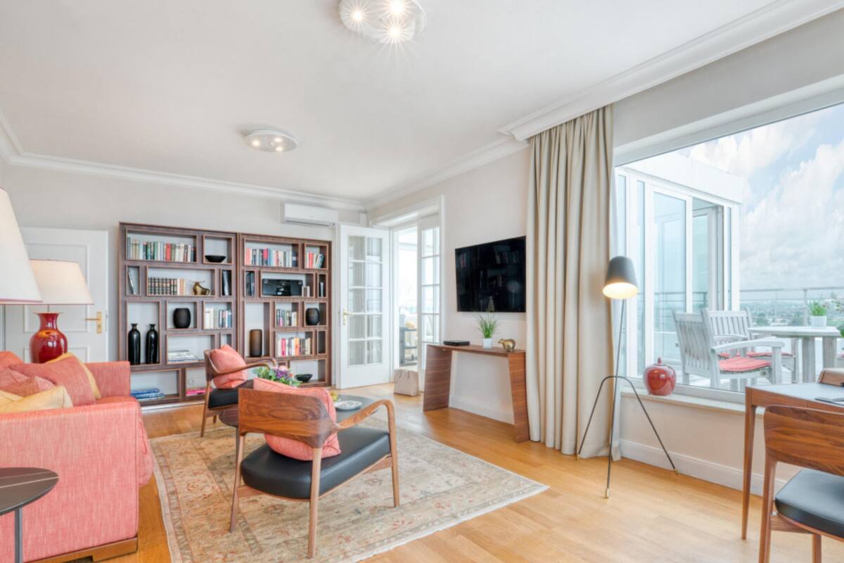 A spacious living room with a wooden floor, a pink sofa, a bookshelf, a television, and a large window with a view.