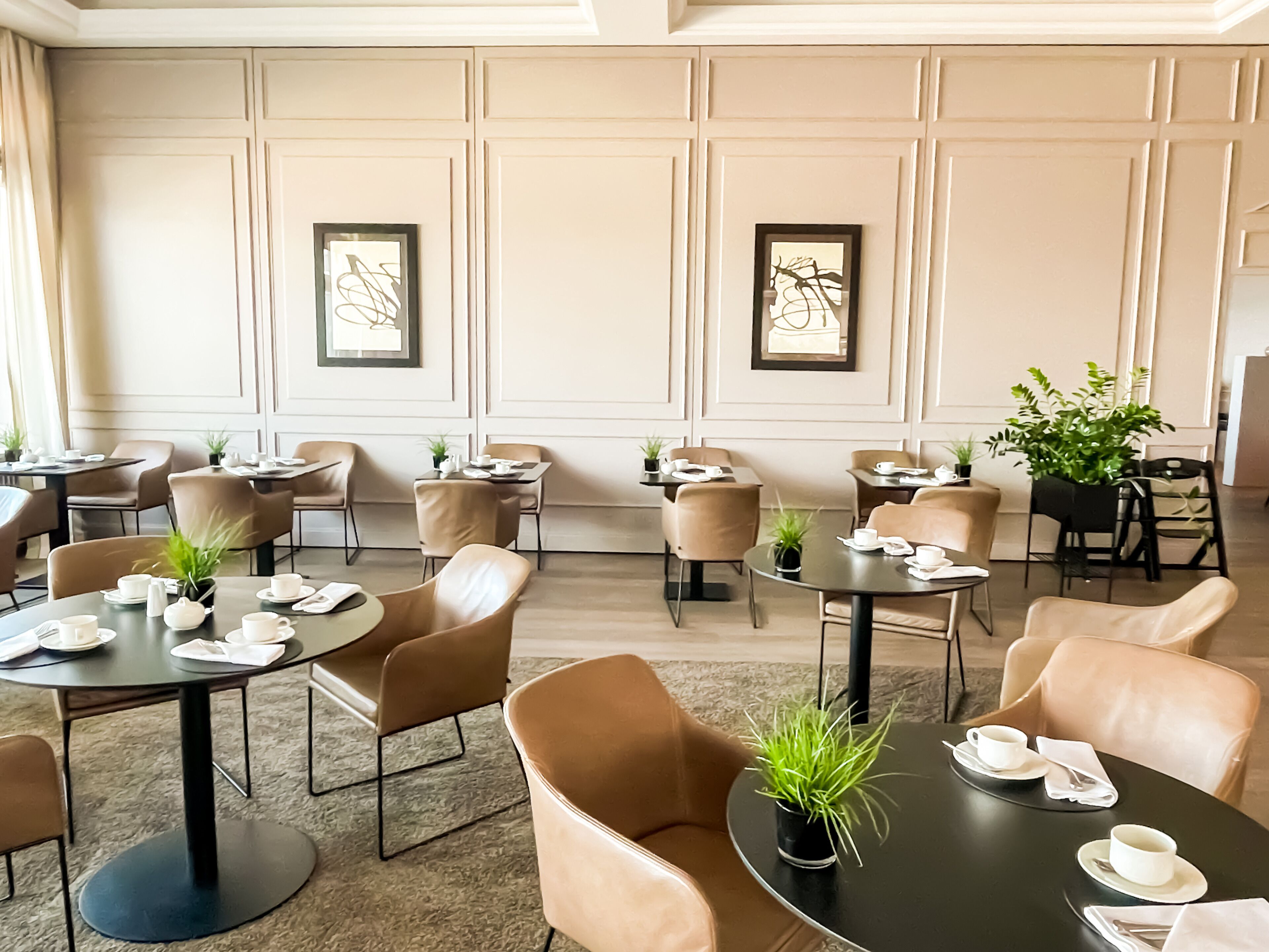Un comedor de hotel con mesas redondas, sillas color beige y plantas en macetas.