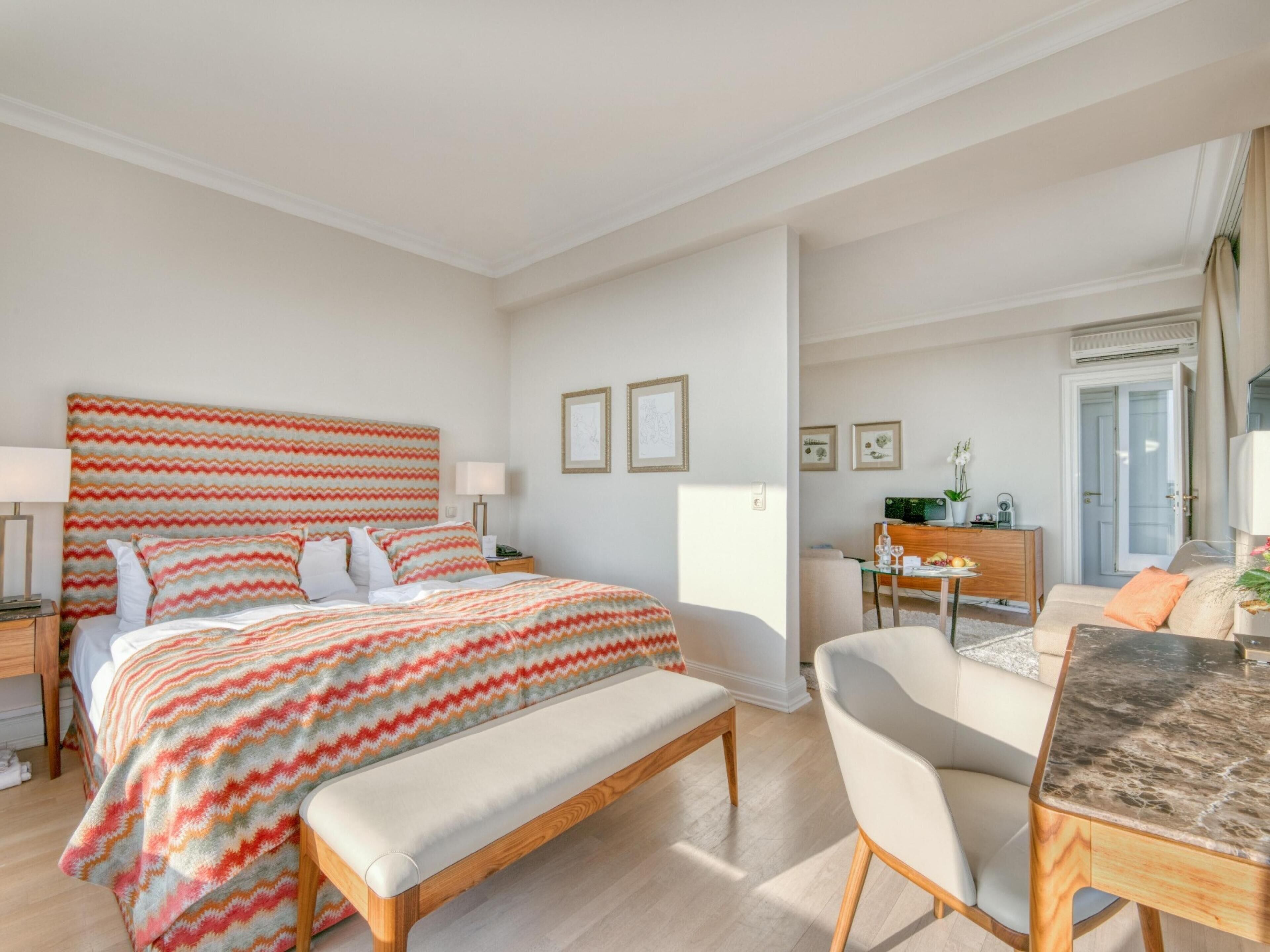 A modern hotel room with a large bed, a wooden bench, a dining table, and a view of the outside.