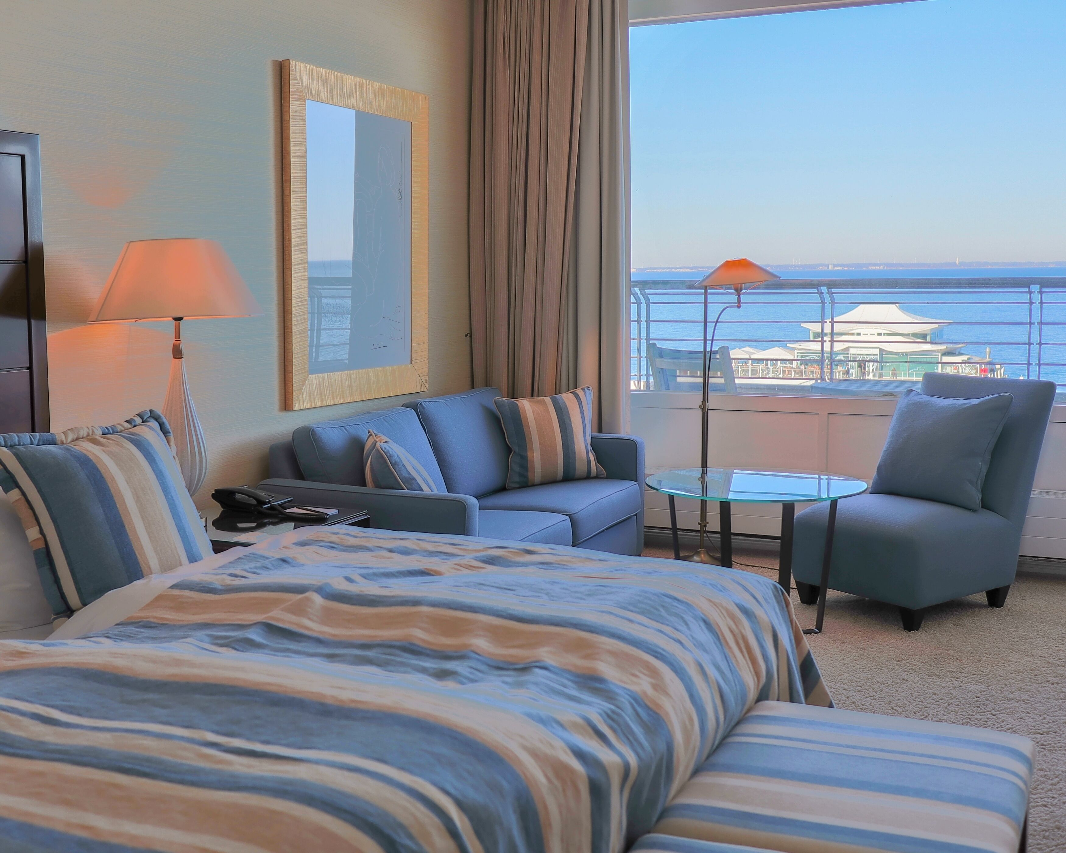 A hotel room with a large bed, a sofa, a chair, a small table, a lamp, and a view of the sea.