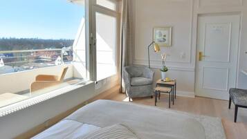 A hotel room with a large bed, a chair, a small table, and a view of a cityscape through a large window.