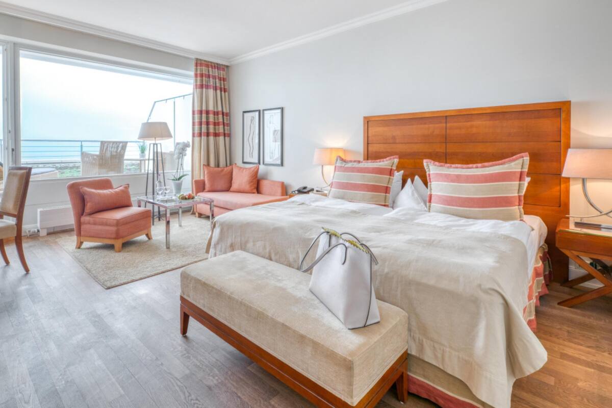 A hotel room with a large bed, a sofa, a chair, a small table, and a view of the sea.