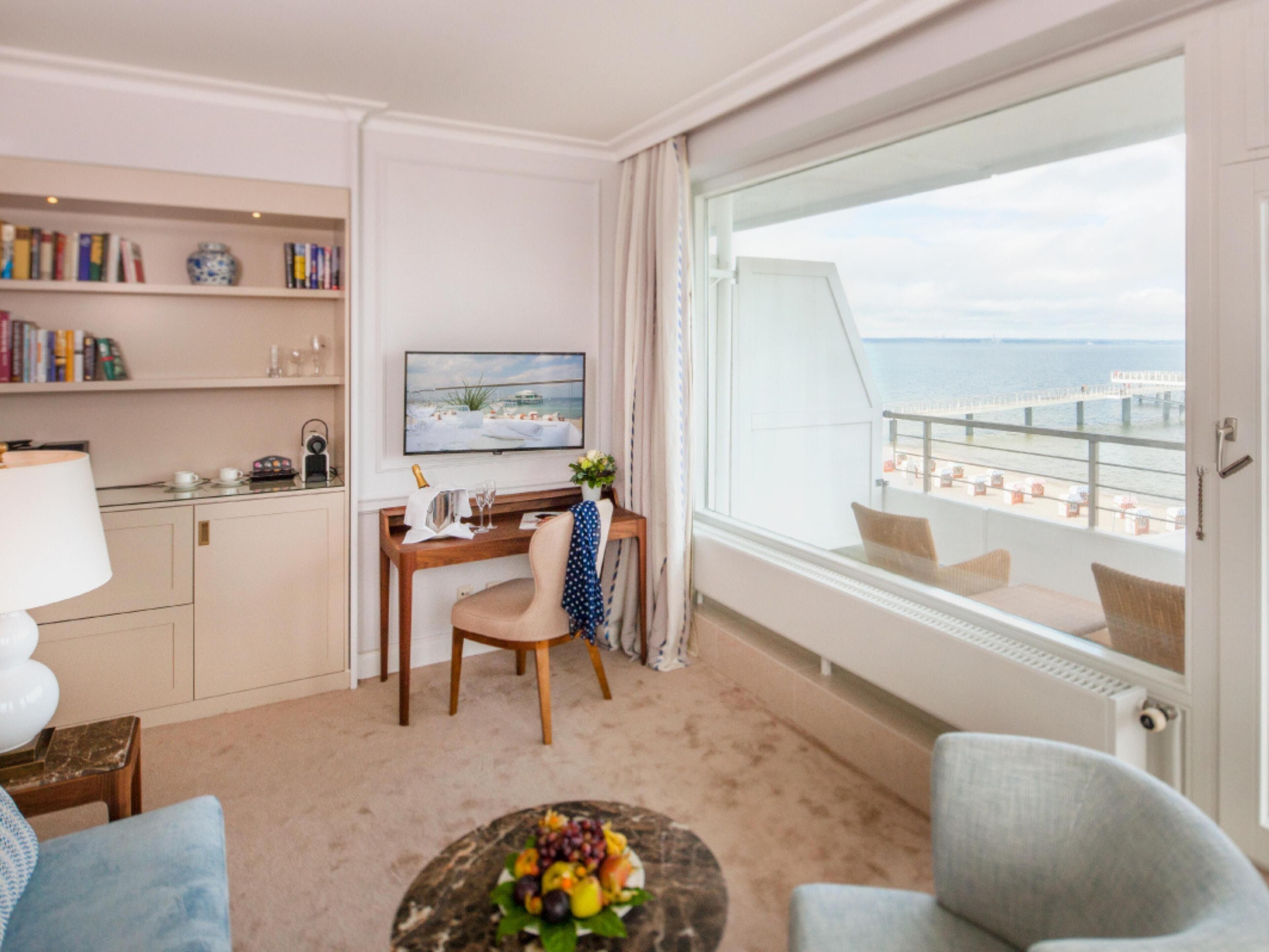 A hotel room with a desk, a chair, a coffee table with fruit, a bookshelf, a television, and a large window with a view of the sea and a pier.