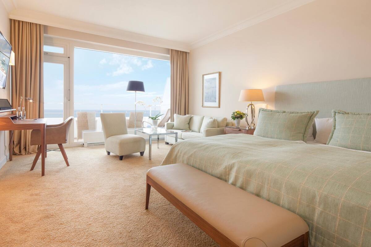 A hotel room with a large bed, a desk, a chair, a sofa, and a bench. There is a window with curtains and a view of the sky.