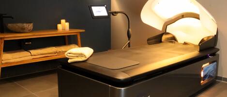 Sala de massagem com mesa preta, iluminação fixada na parede e uma tela pequena exibindo um vídeo.