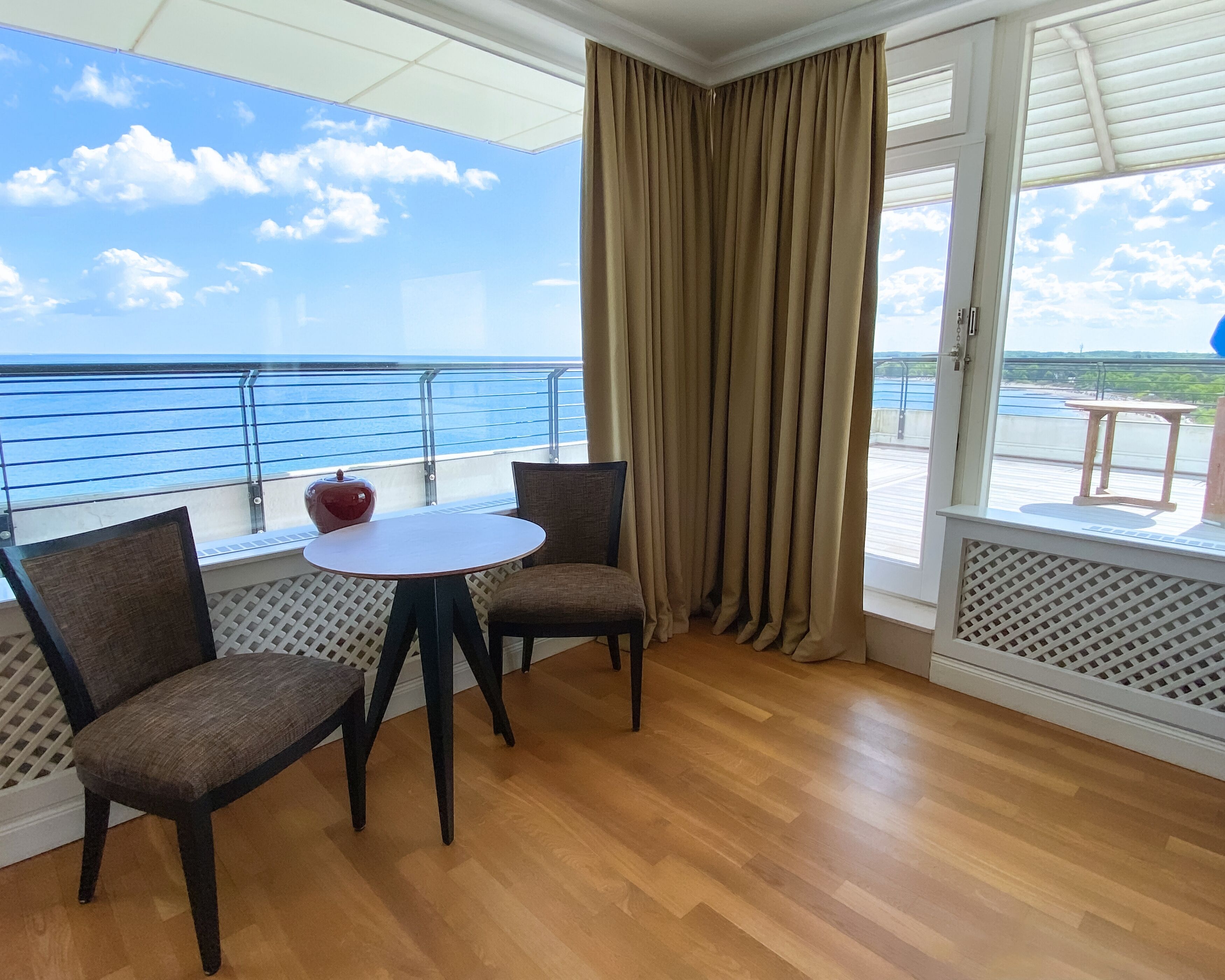 Een balkon met een houten vloer, twee stoelen, een klein rond tafeltje en uitzicht op zee.