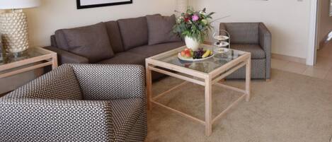 Una sala de estar con un sofá, dos sillones, una mesa de centro con fruta y dos lámparas.