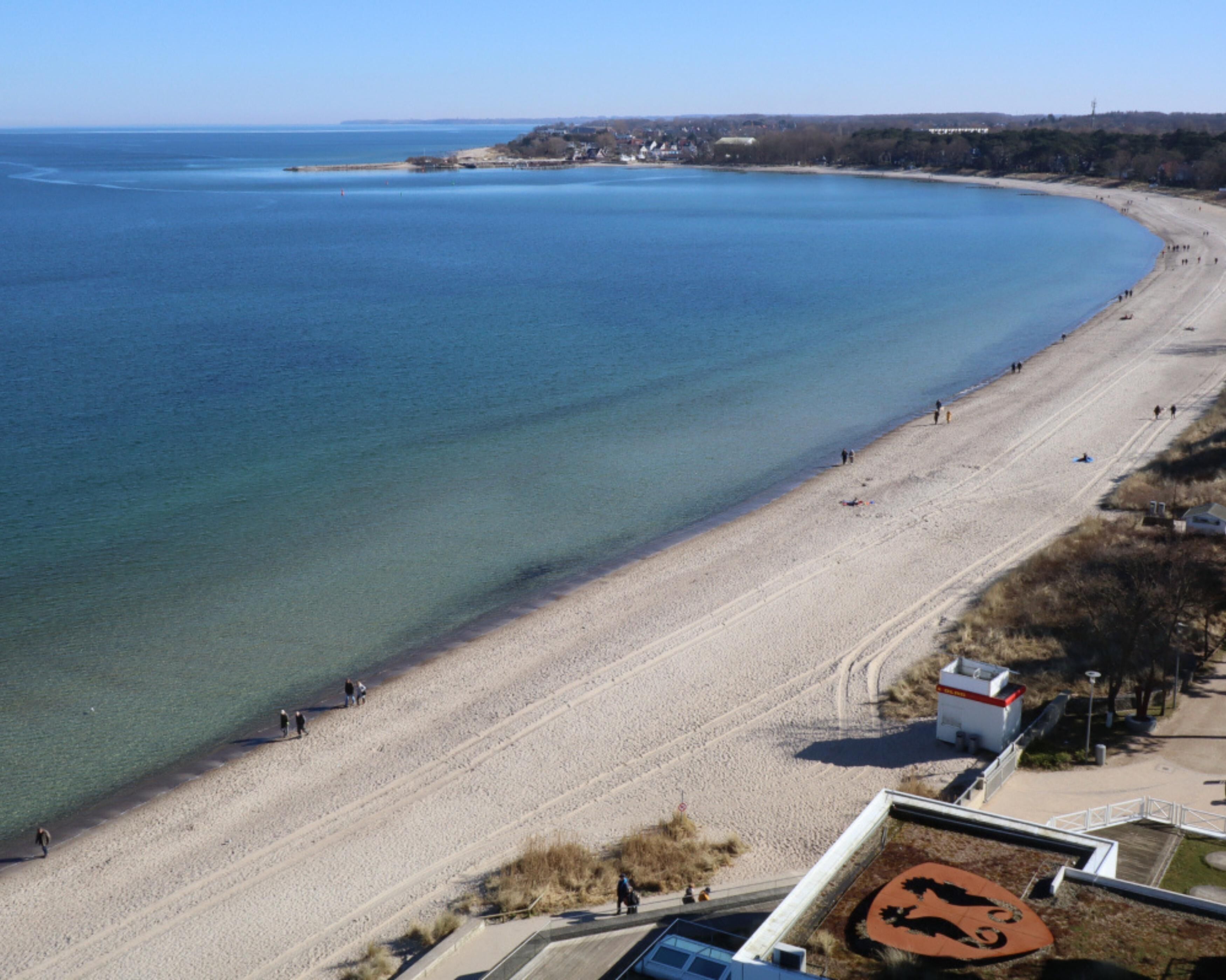 An aerial view of a beach with clear blue water, a sandy shore, and a few people walking along the beach.