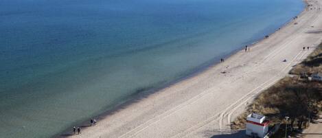 An aerial view of a beach with clear blue water, a sandy shore, and a few people walking along the beach.