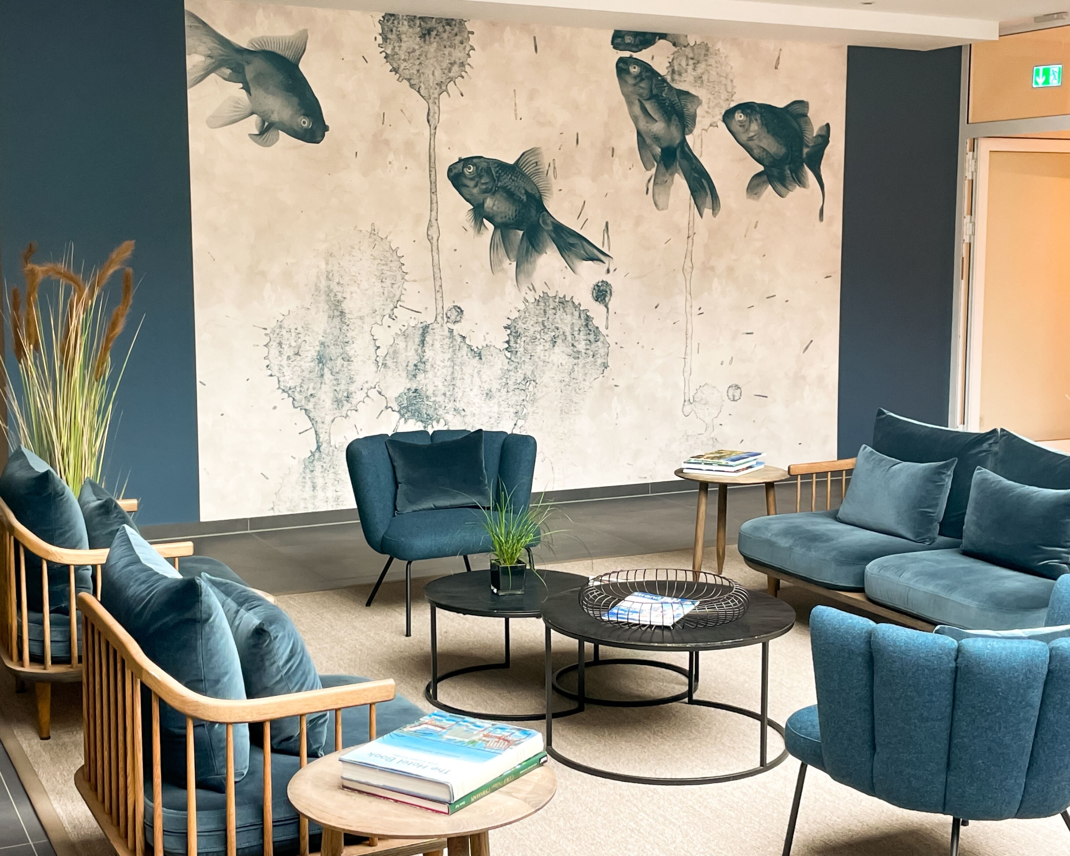 A modern lounge area with blue upholstered chairs, a round coffee table, and a wall mural of fish.