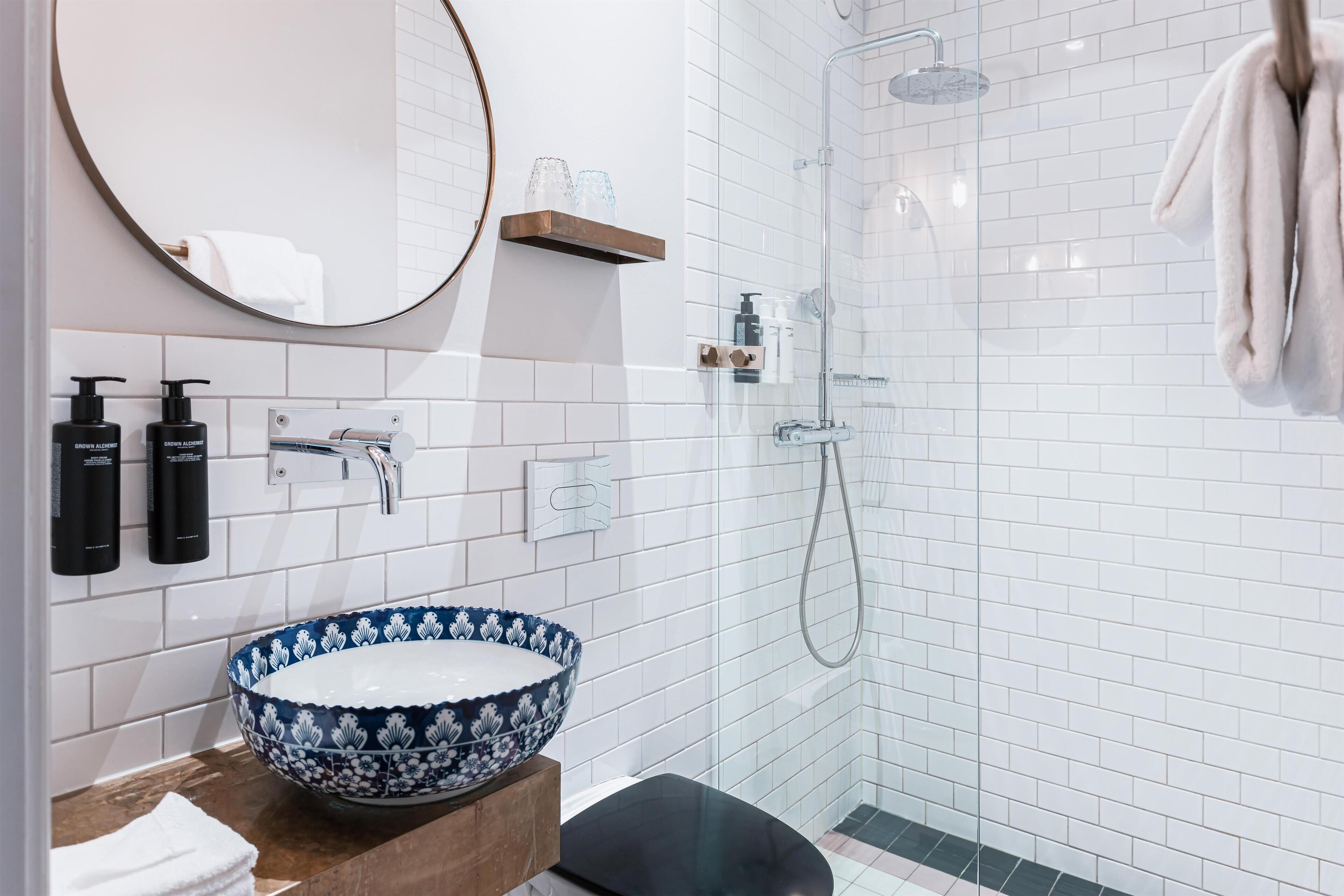 A modern bathroom with a round mirror, a blue and white patterned basin, a shower with a glass door, and a towel hanging on the wall.
