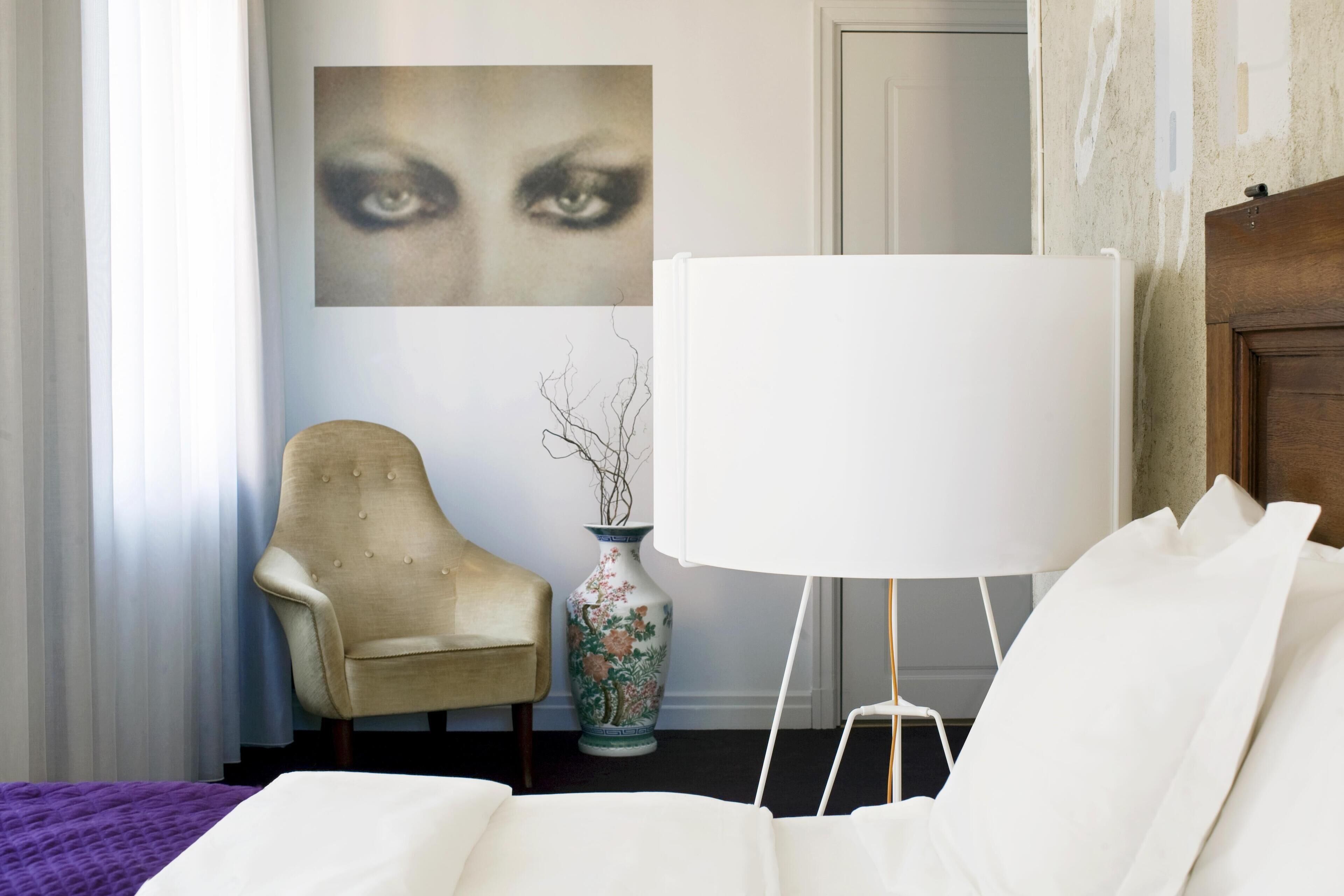 A hotel room with a bed, a chair, a vase, and a wall with a painting.