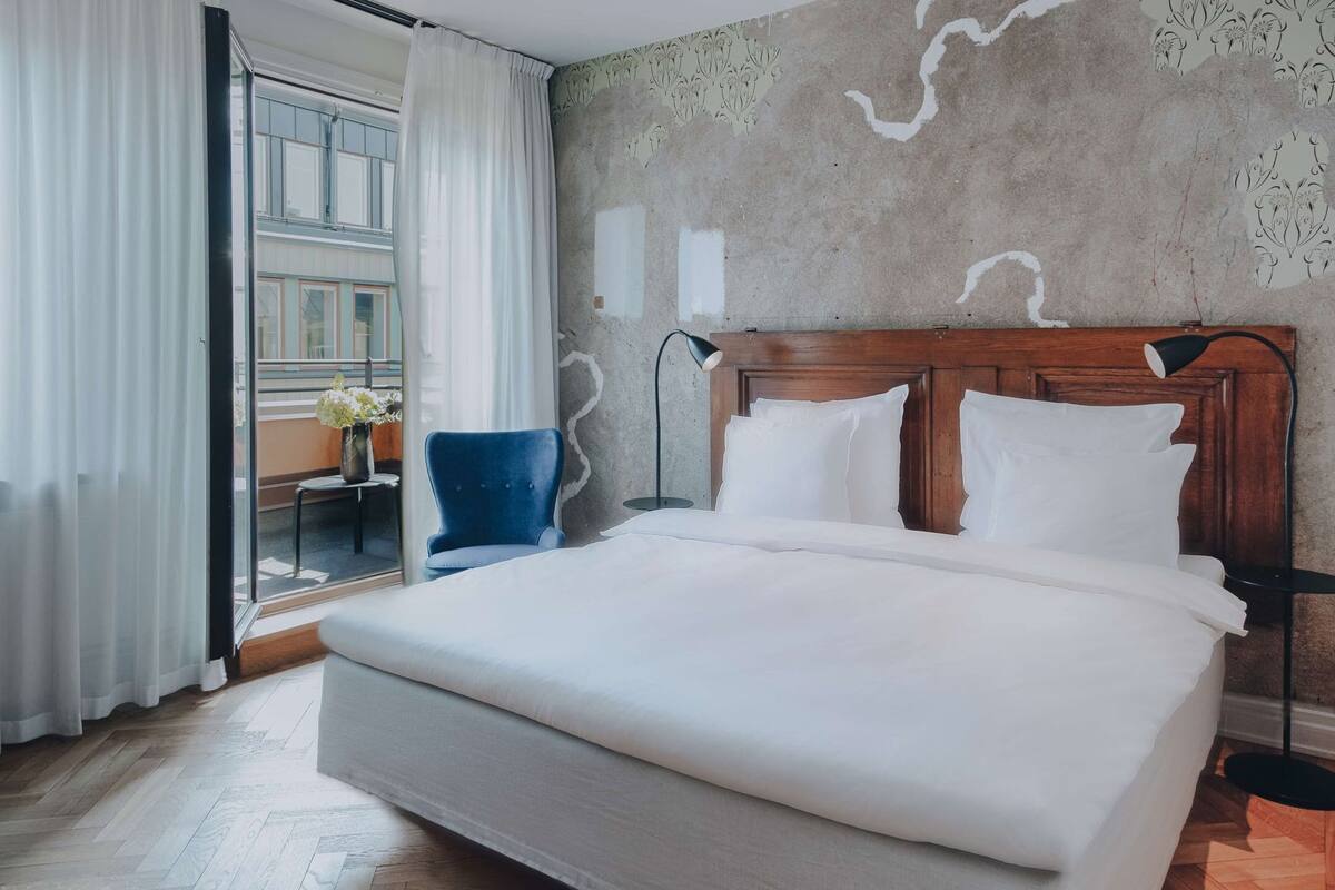 A large bed with white linens, a blue chair, and a view through a glass door.