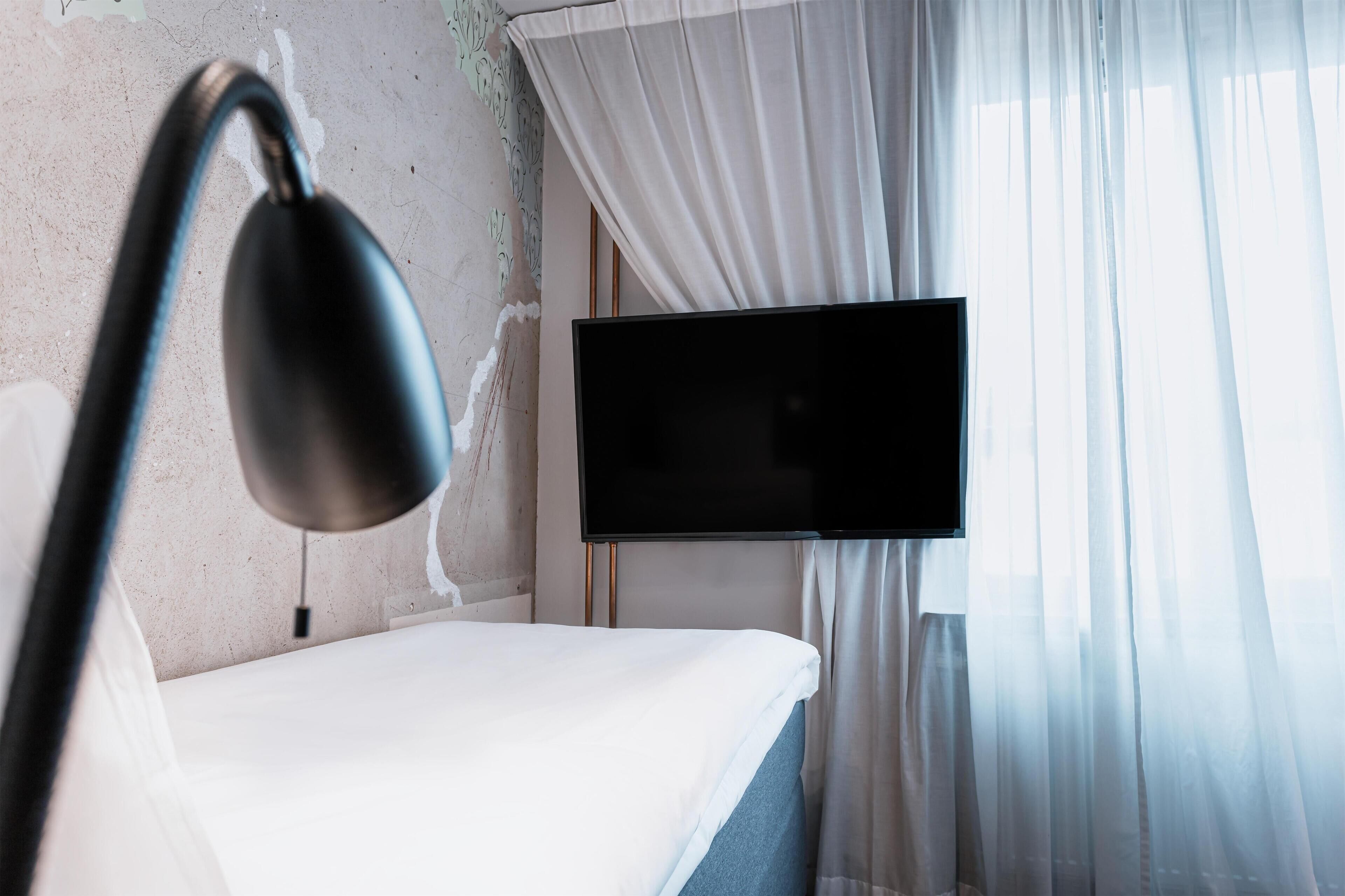 A hotel room with a bed, a flat-screen TV mounted on the wall, and sheer curtains.