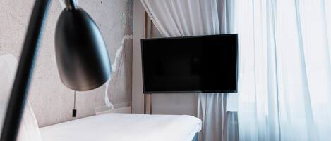 A hotel room with a bed, a flat-screen TV mounted on the wall, and sheer curtains.