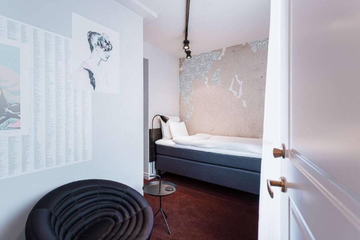 A room with a bed, a wall-mounted lamp, a small round table, and a black chair.