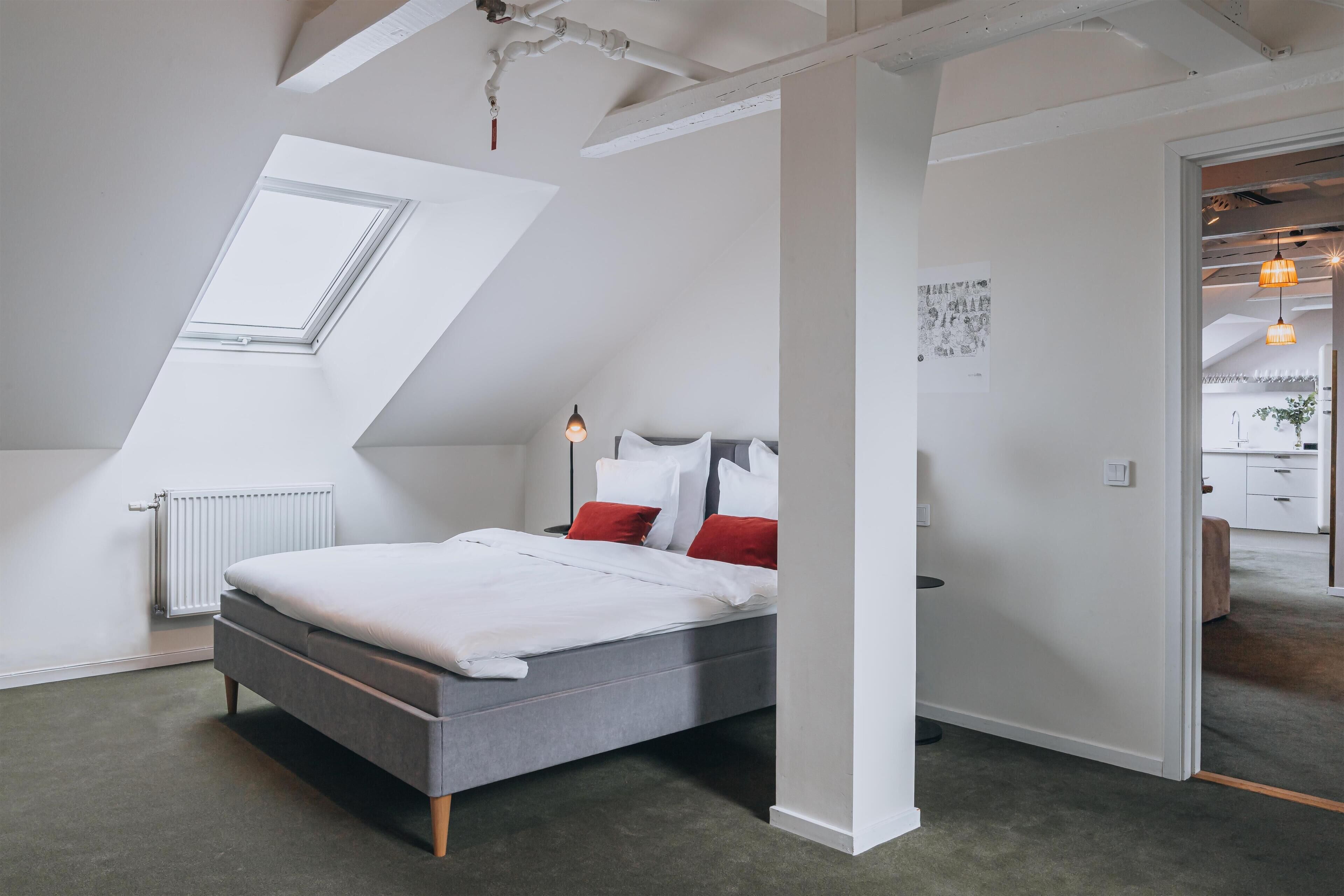 A modern bedroom with a sloped ceiling, a skylight, a bed with white bedding and red pillows, and a radiator.