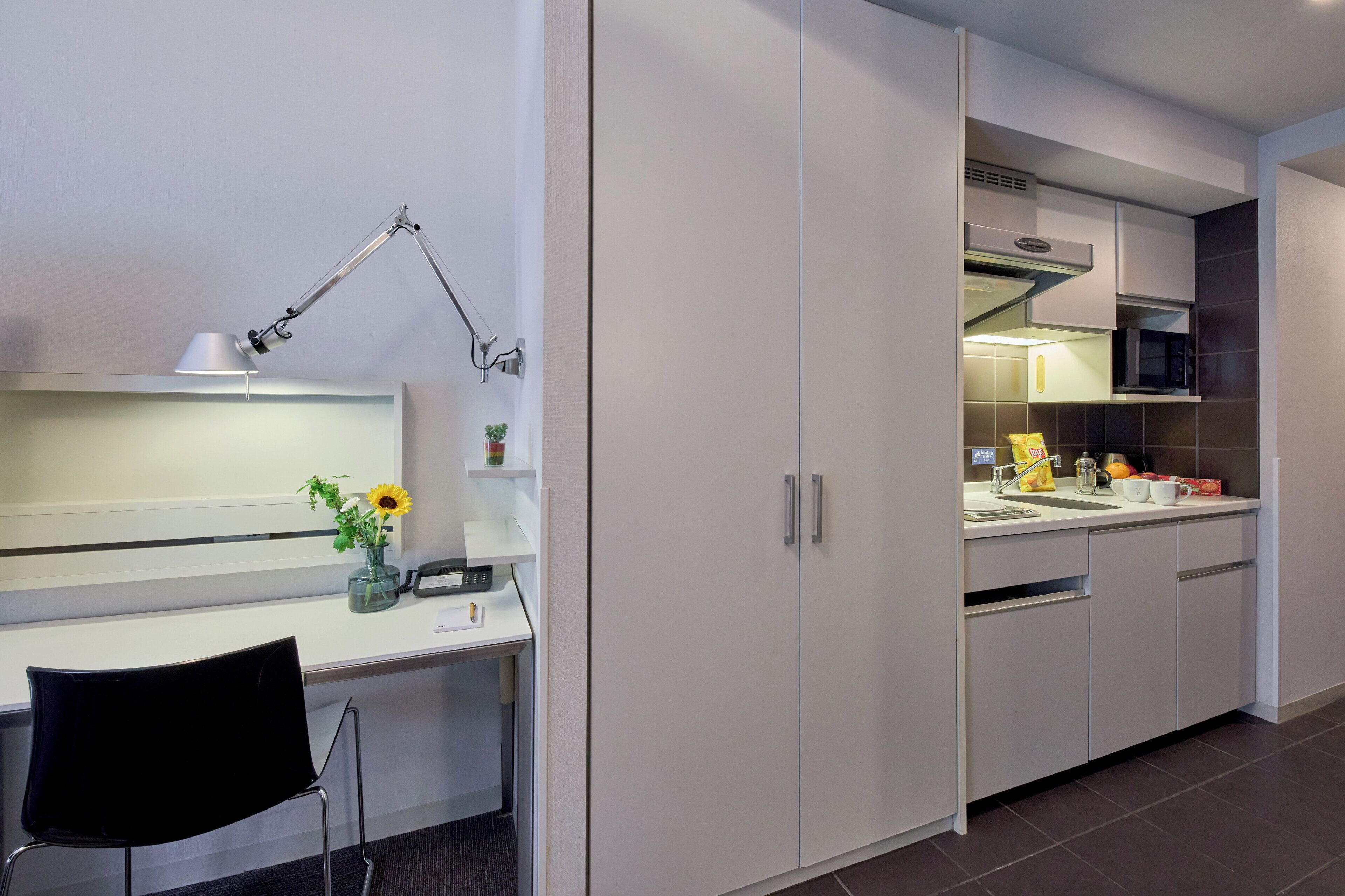 A modern kitchen with white cabinets, a microwave, and a sink. There is a desk with a lamp and a chair.