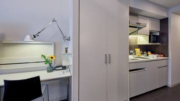 A modern kitchen with white cabinets, a microwave, and a sink. There is a desk with a lamp and a chair.
