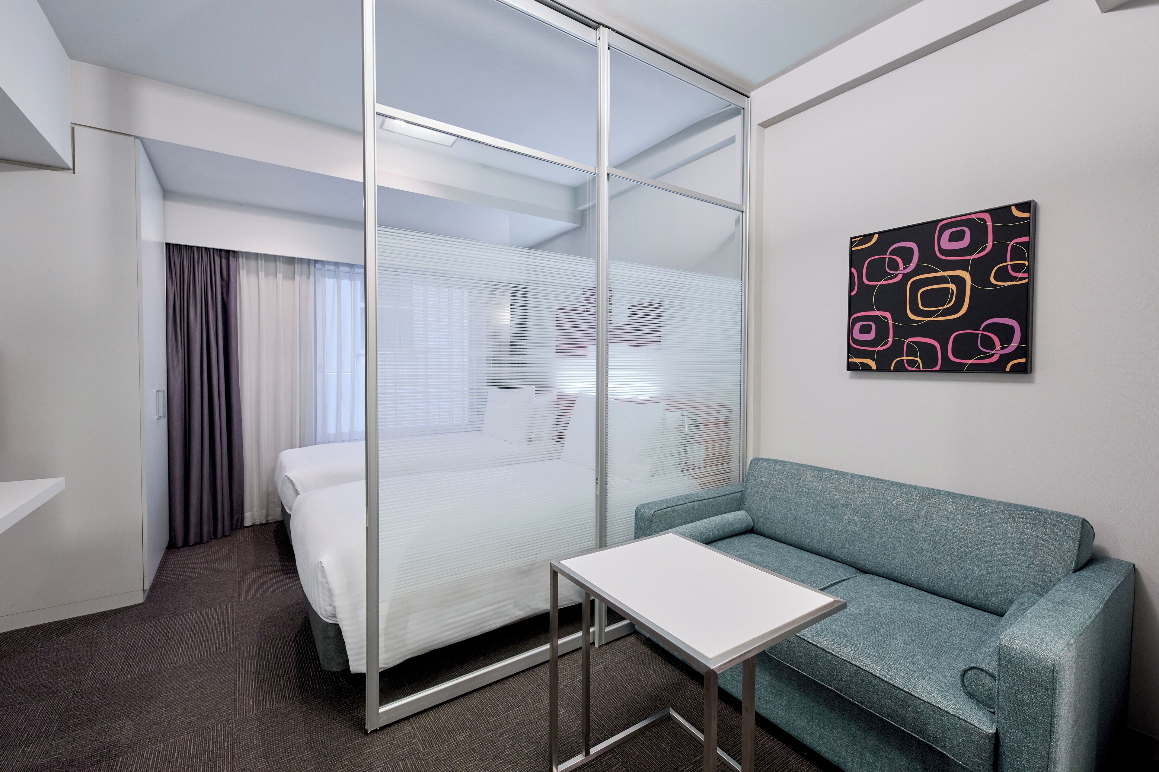 A hotel room with a bed, a sofa, a small table, and a wall-mounted artwork.