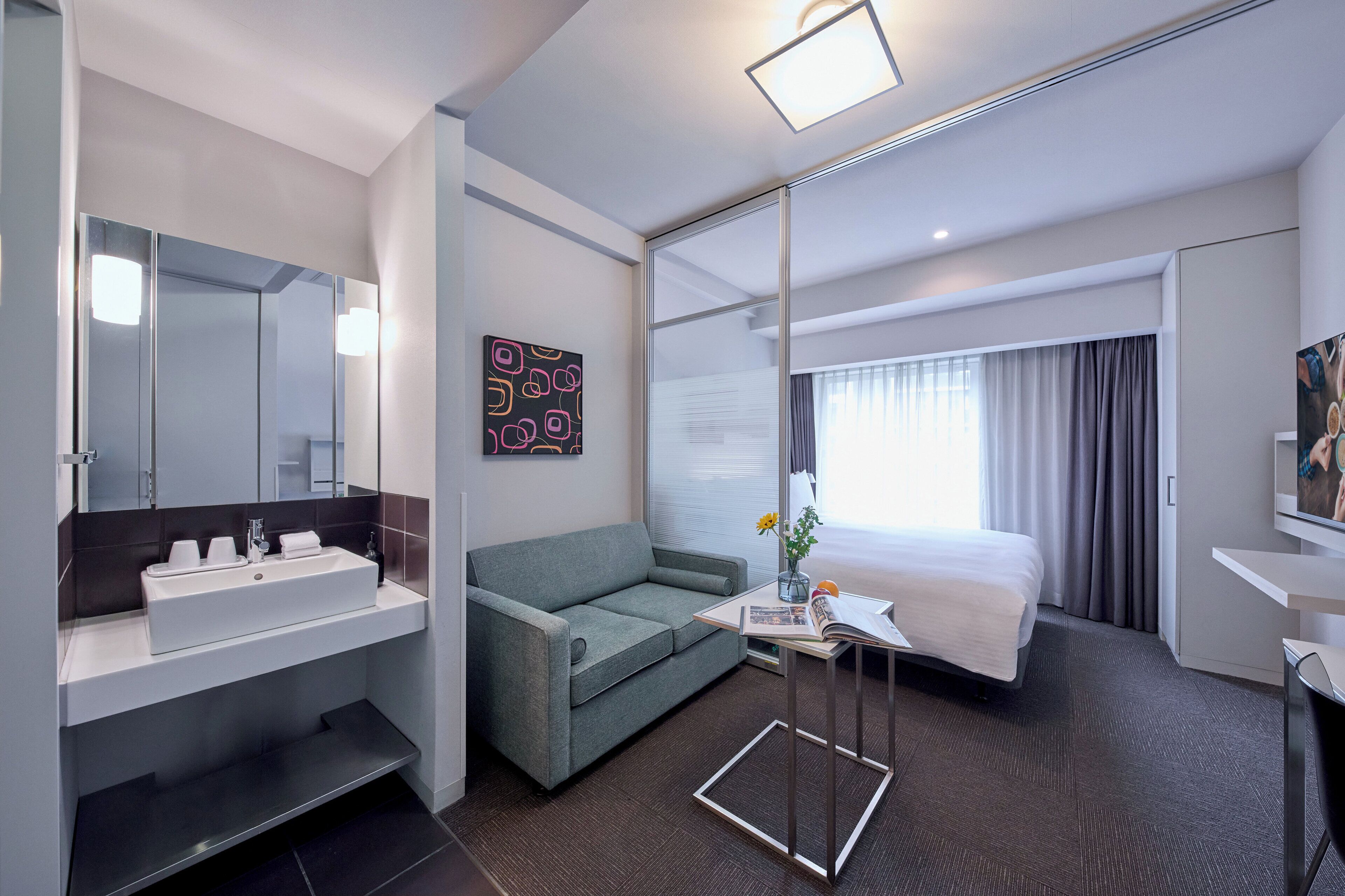 Ein modernes Hotelzimmer mit einem großen Bett, einer Sitzecke, einem Schreibtisch und einem Badezimmer mit Waschbecken und Spiegel.