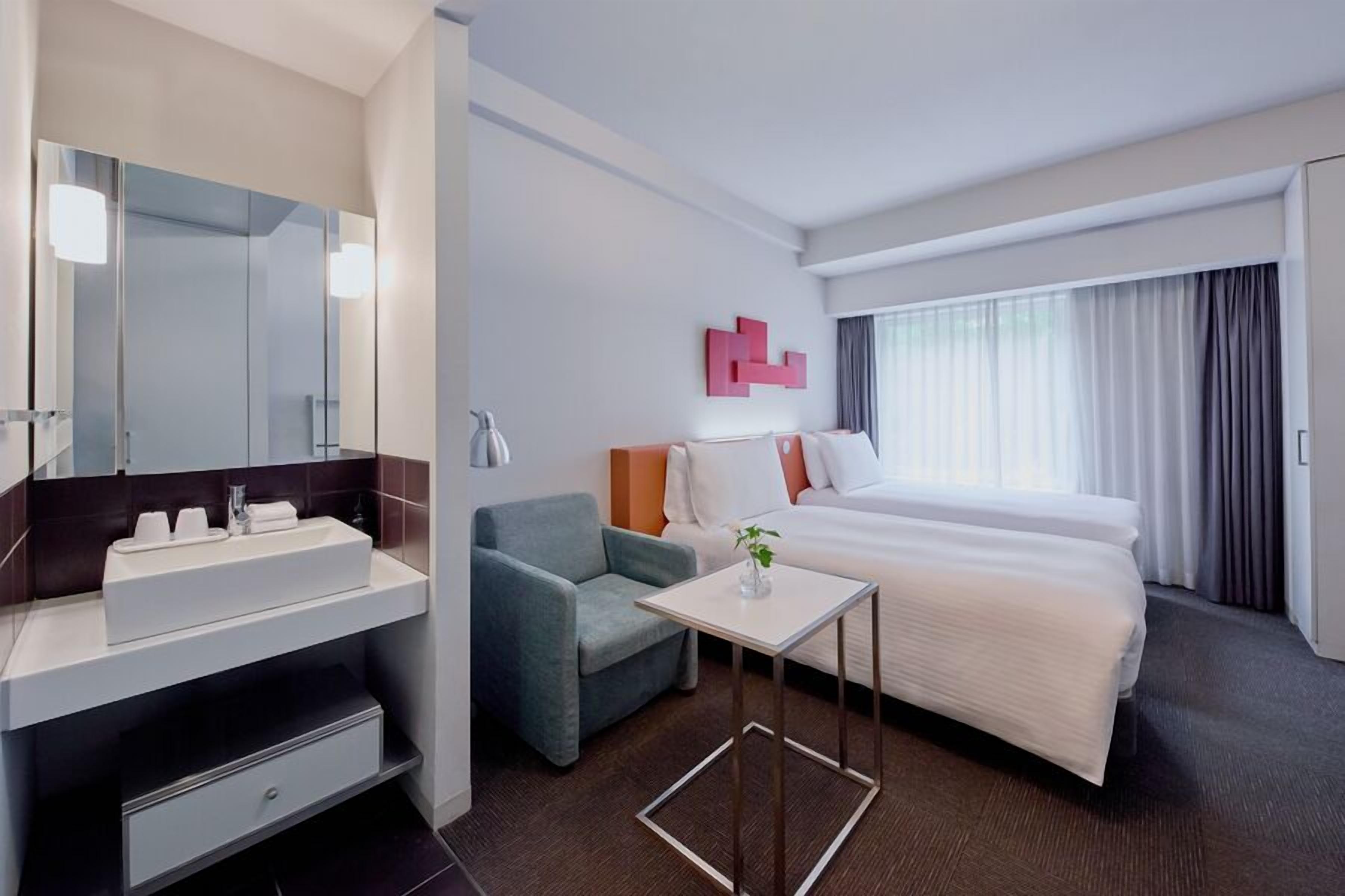 A hotel room with a bed, a bedside table, a chair, a mirror, a sink, and a window with curtains.