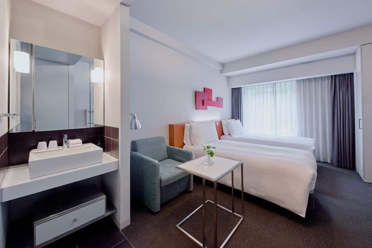 Ein Hotelzimmer mit Bett, Nachttisch, Sessel, Spiegel, Waschbecken und einem Fenster mit Vorhängen.